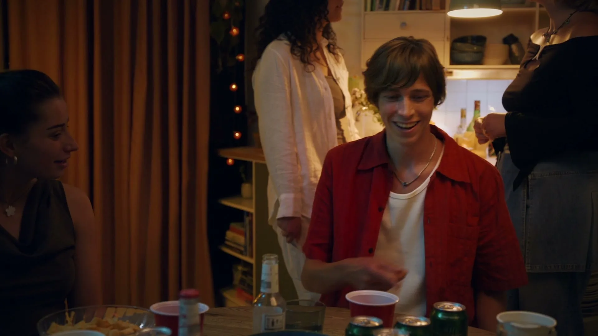 A young man in a red shirt smiles and talks to a young woman at a lively house party, with other guests visible in the background.
