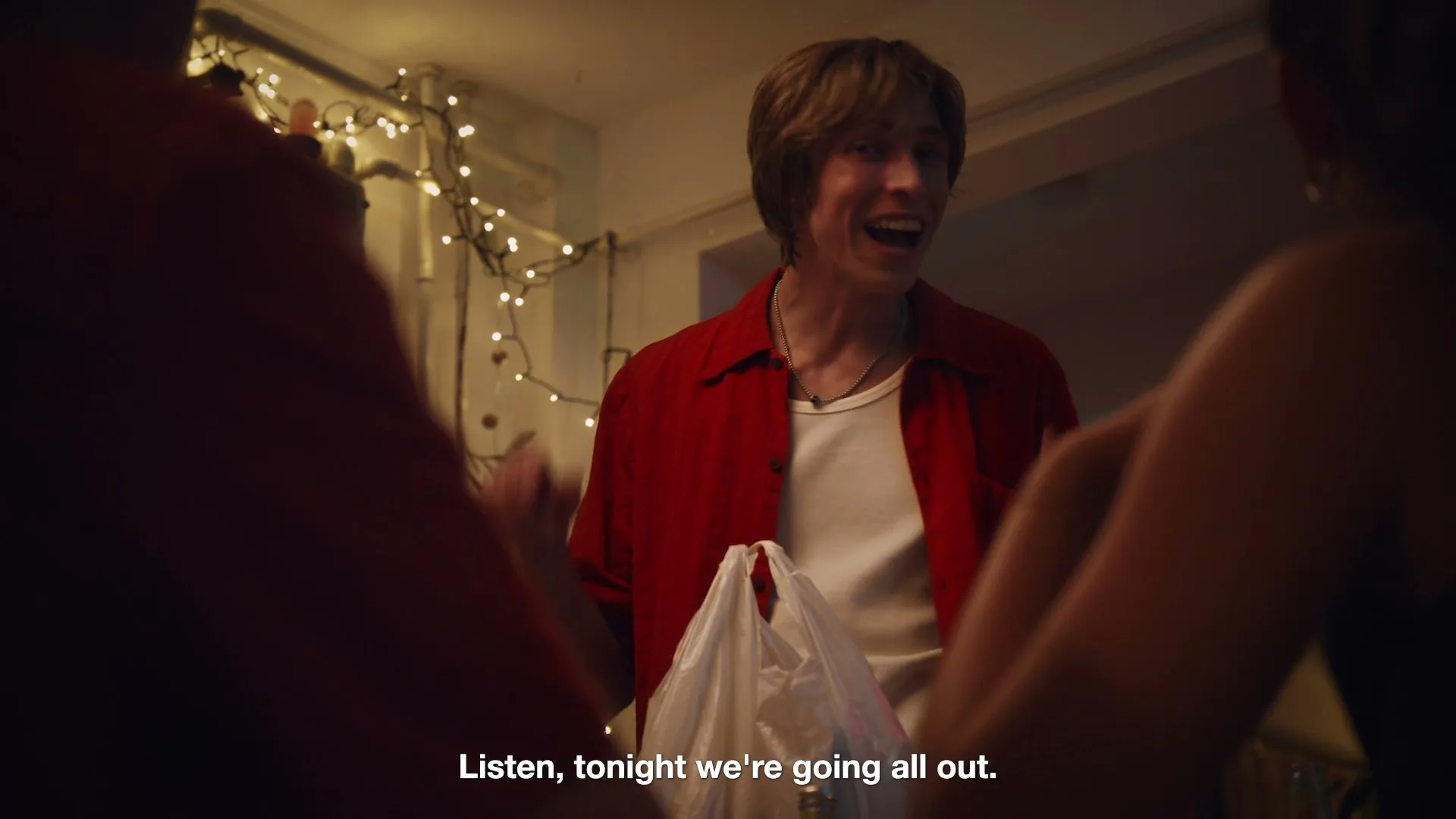 A young man with shaggy hair and a red shirt smiles, holding a plastic bag, as he talks to two people at a party entrance.