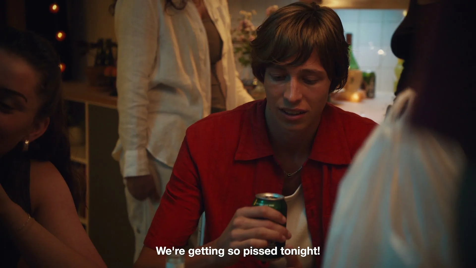 A young man in a red shirt holds a beer can, looking down, while a young woman next to him laughs at a lively house party.
