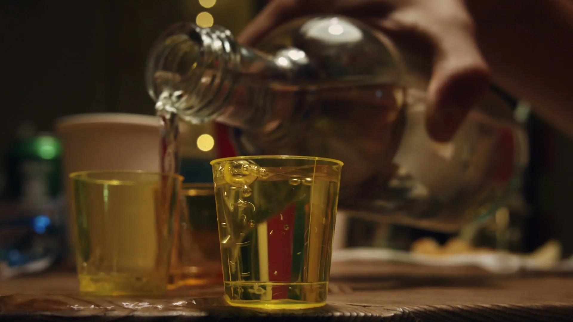 A person's hand pours clear liquid from a bottle into a small yellow plastic cup on a wooden table at a party.