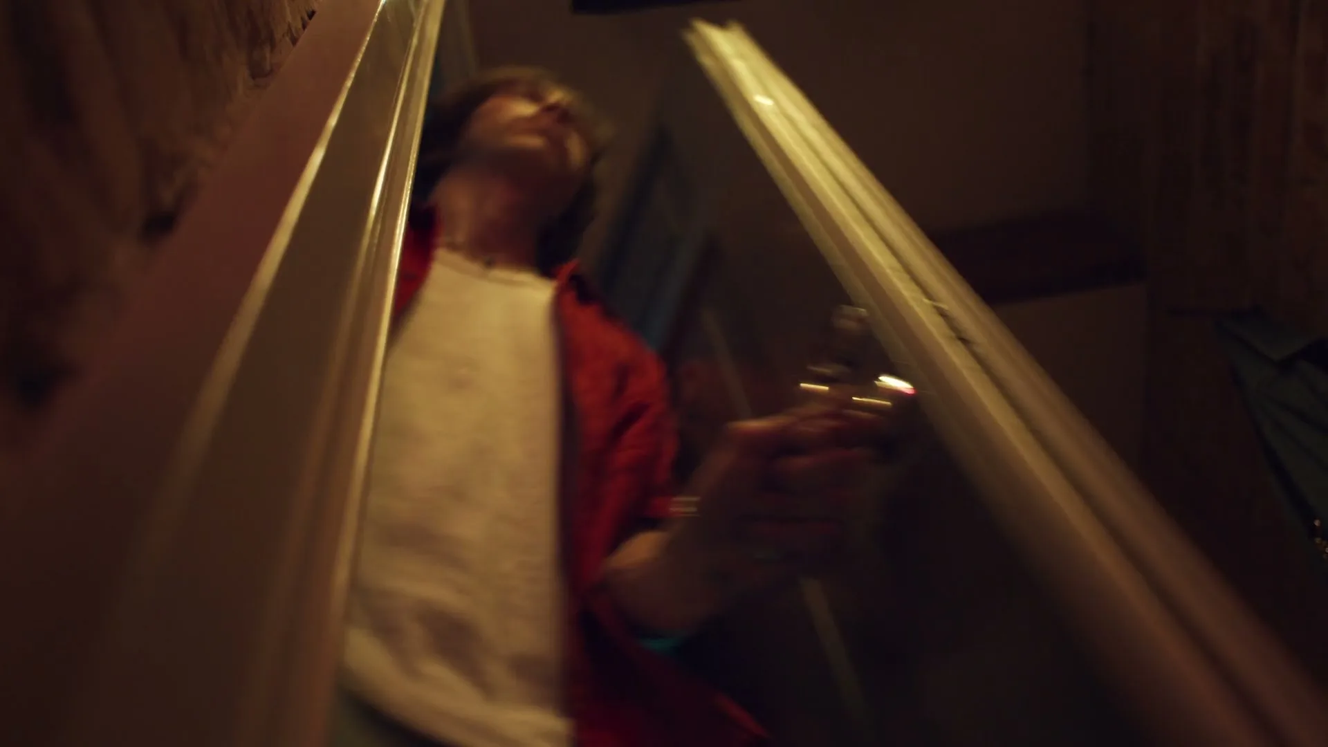 A man in a red shirt and white t-shirt opens a door and walks out of a dark room, seen from a low angle.