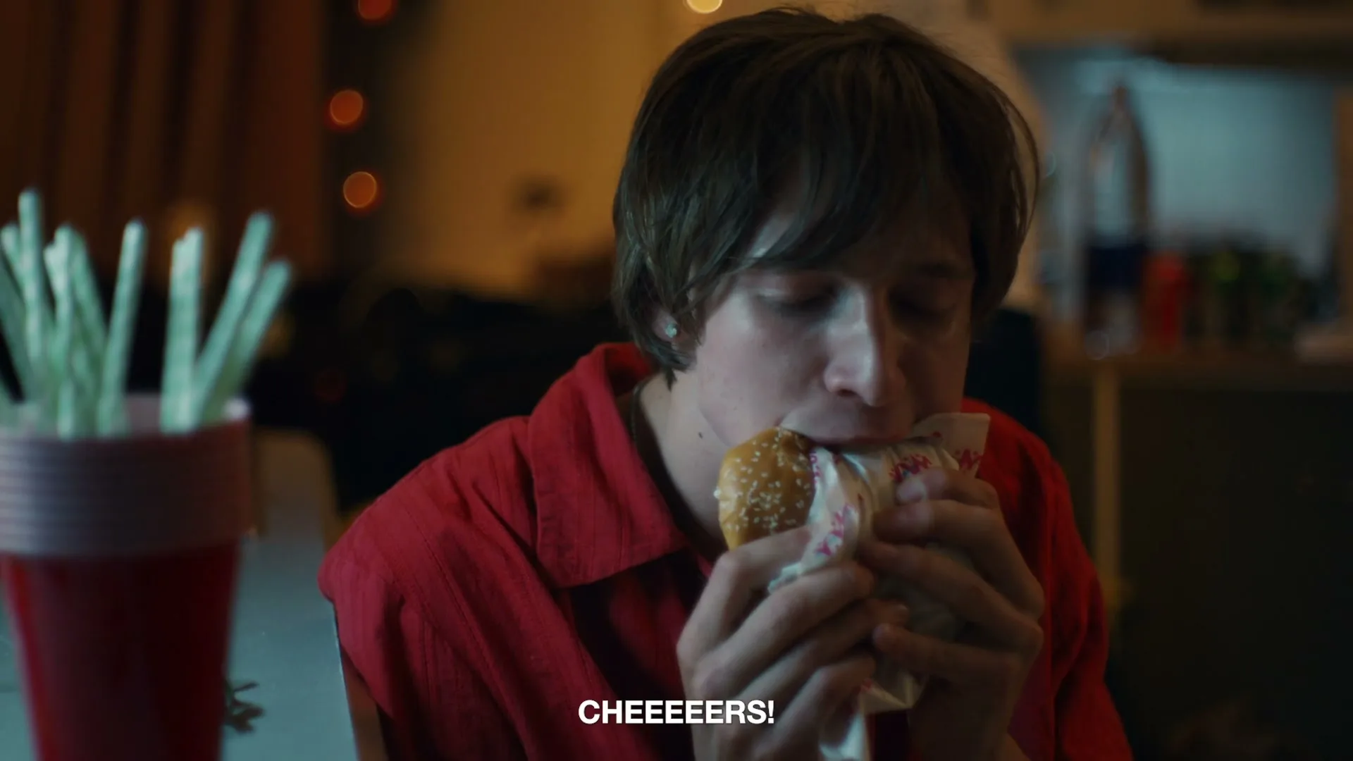 A young man in a red shirt devours a burger, with food spilling from his mouth, at a dimly lit party.