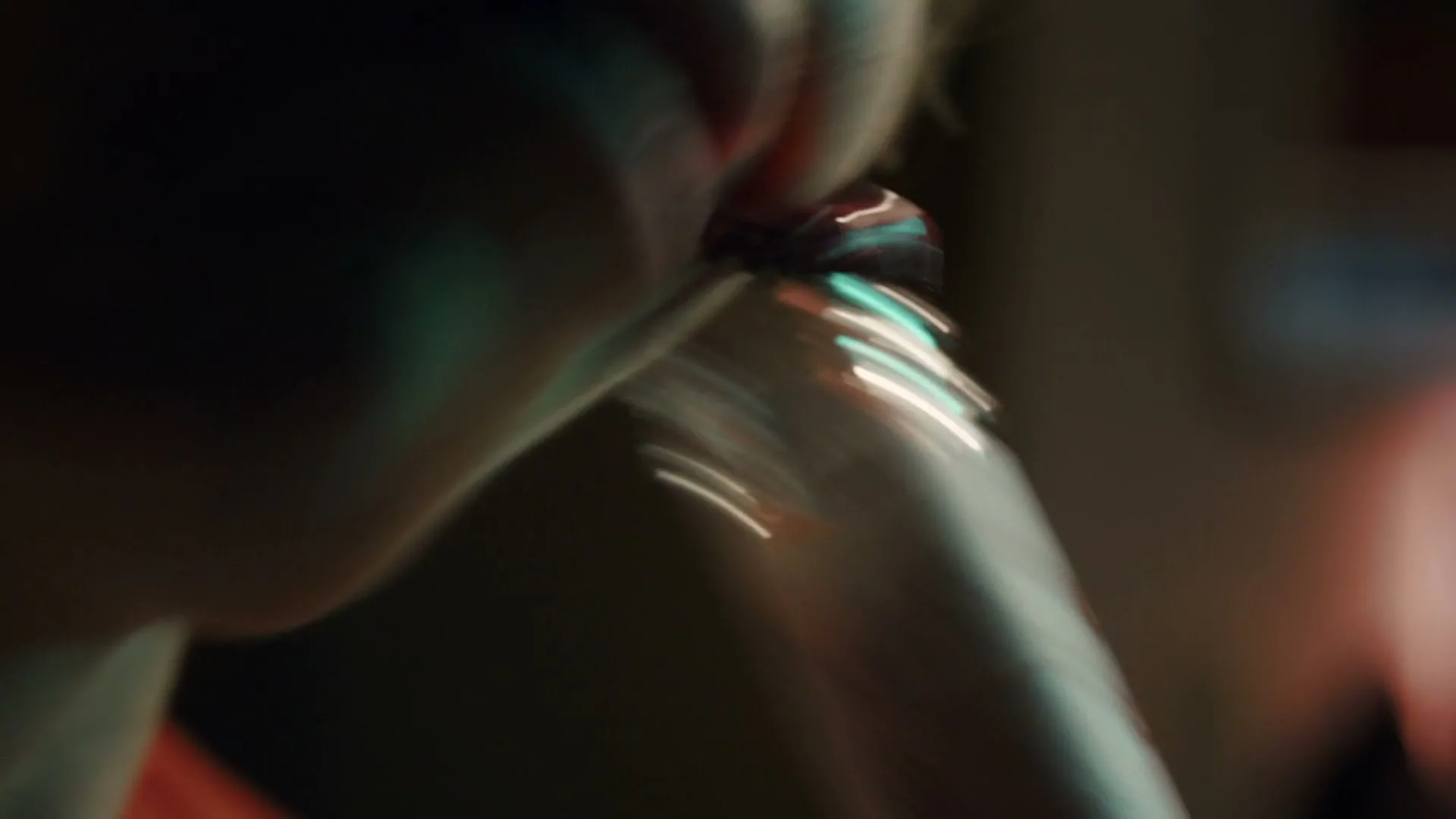 A person's mouth is seen in extreme closeup as they drink from a bottle, with green and red light reflecting on their face and the bottle.