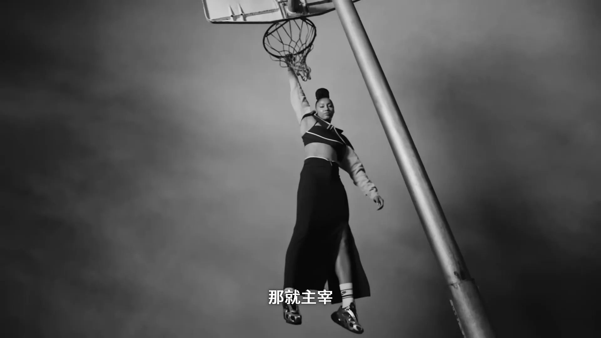 A young woman in athletic wear and a long skirt hangs from a basketball hoop, looking determined in a black and white shot.