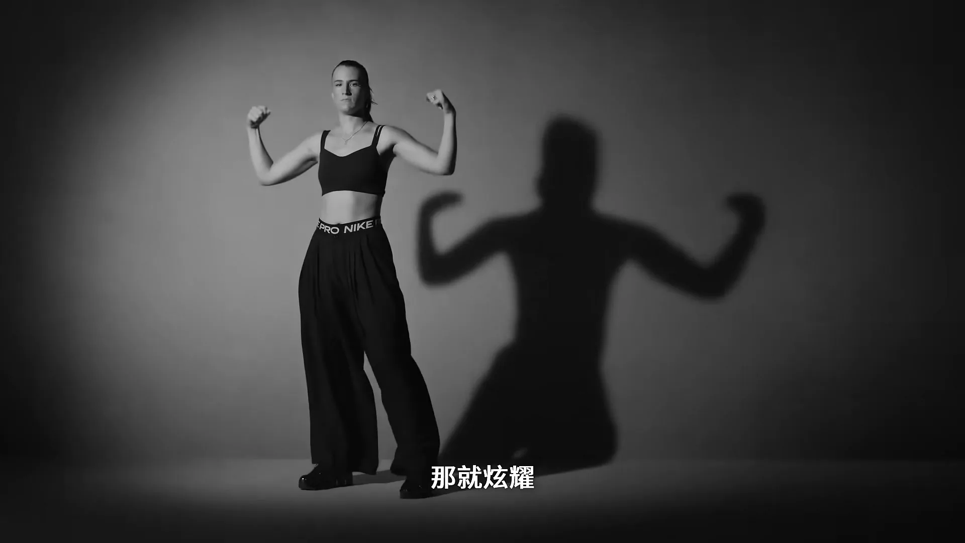 A female athlete in a sports bra and loose pants flexes her biceps, casting a large shadow on a plain grey background.