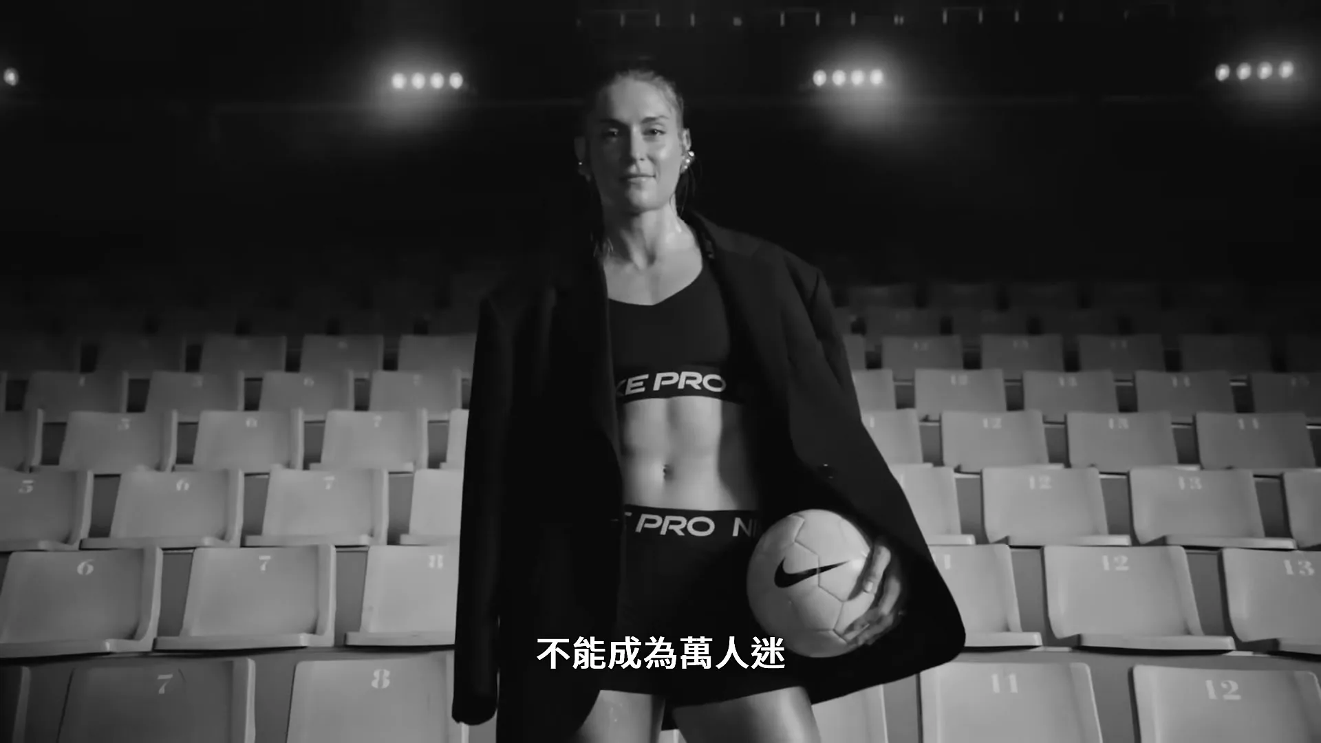 A female athlete in a sports bra and shorts, with an oversized blazer, holds a soccer ball in a stadium, looking directly at the camera.