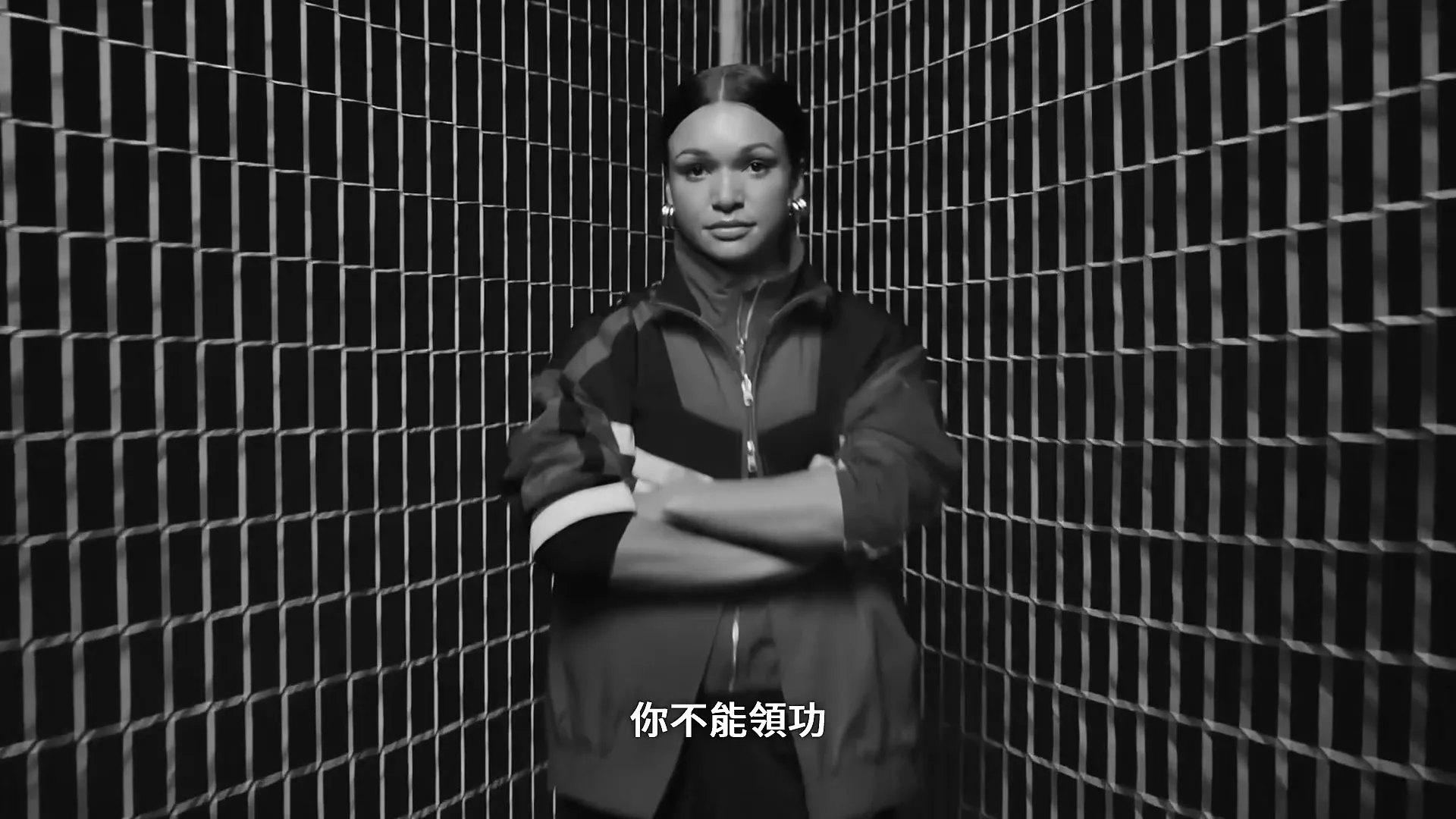 A female athlete in sportswear stands with arms crossed, looking directly at the camera, surrounded by a grid-like structure, in a black and white shot.