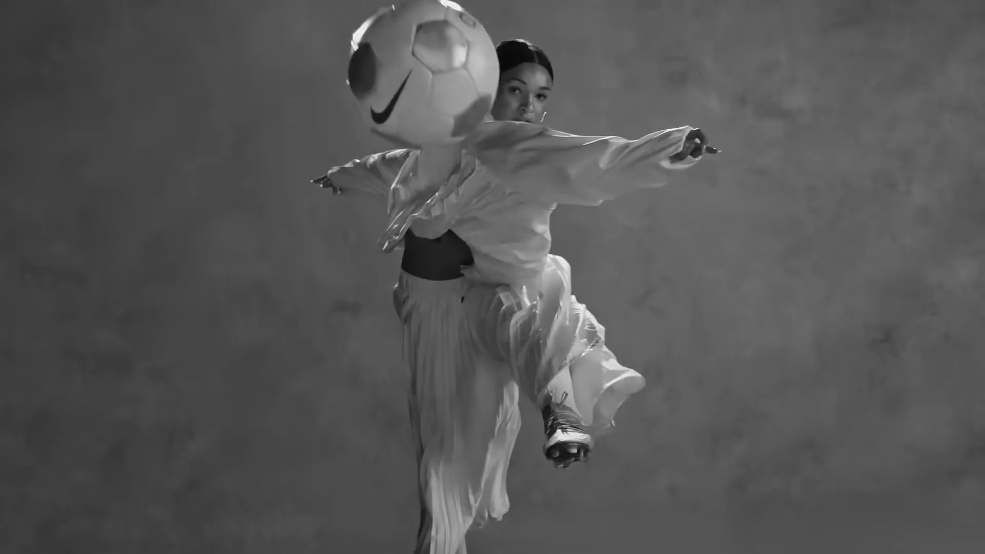 A female soccer player in white athletic wear kicks a soccer ball in mid-air against a smoky grey background.