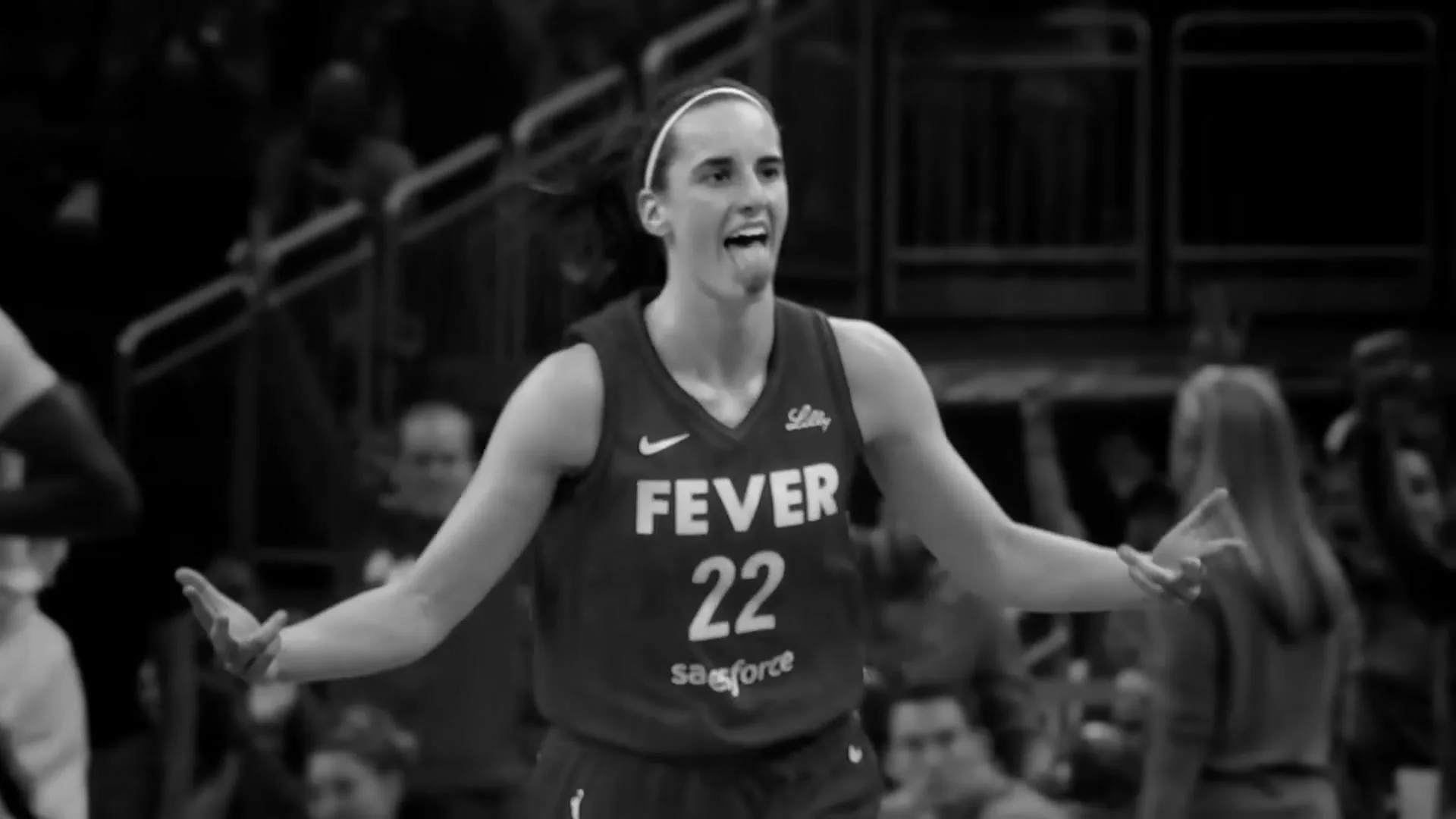 A female basketball player in a "Fever 22" jersey celebrates with arms outstretched and tongue out on a basketball court.