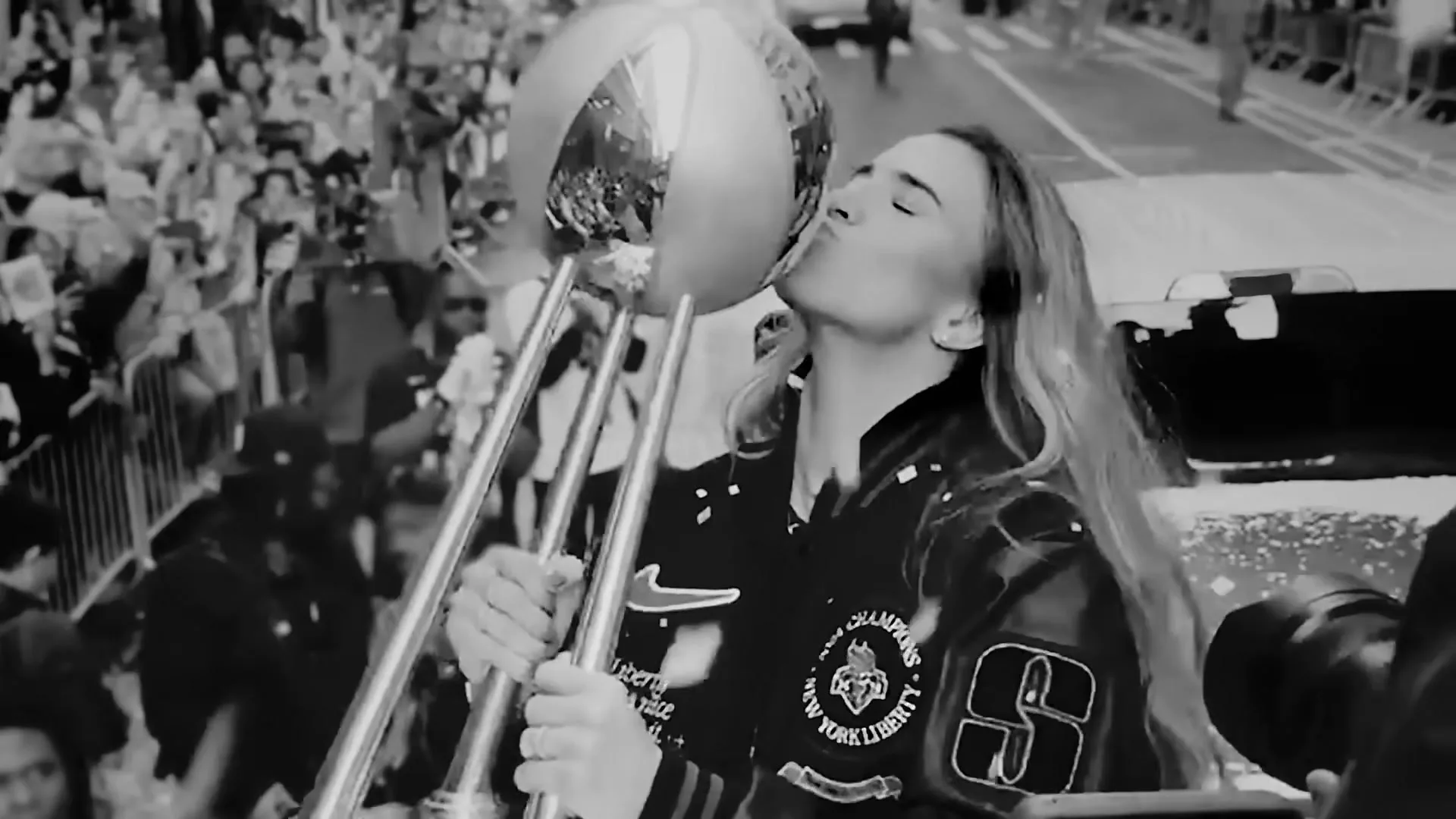 A female athlete in a varsity jacket kisses a large, shiny trophy during a parade, with a crowd visible in the background.