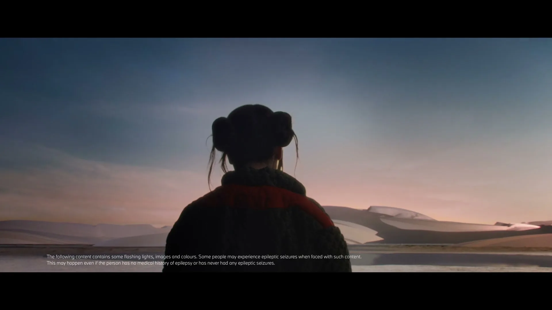 A woman with hair buns and a chunky sweater stands with her back to the camera, looking out at a vast, desolate landscape with sand dunes under a twilight sky.