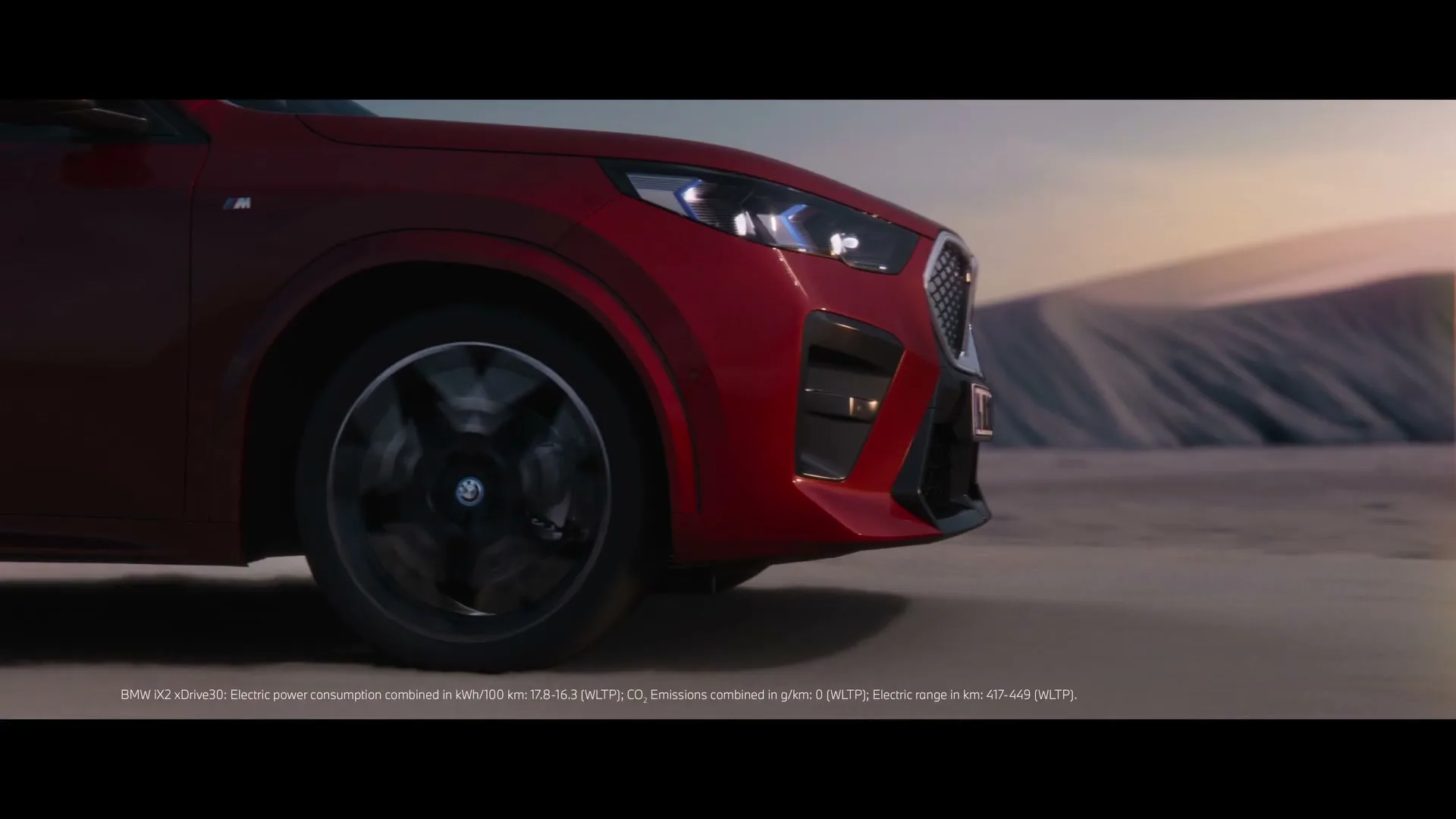 A red BMW car is driving on a desert road, with its front wheel and headlight visible, under a clear sky.
