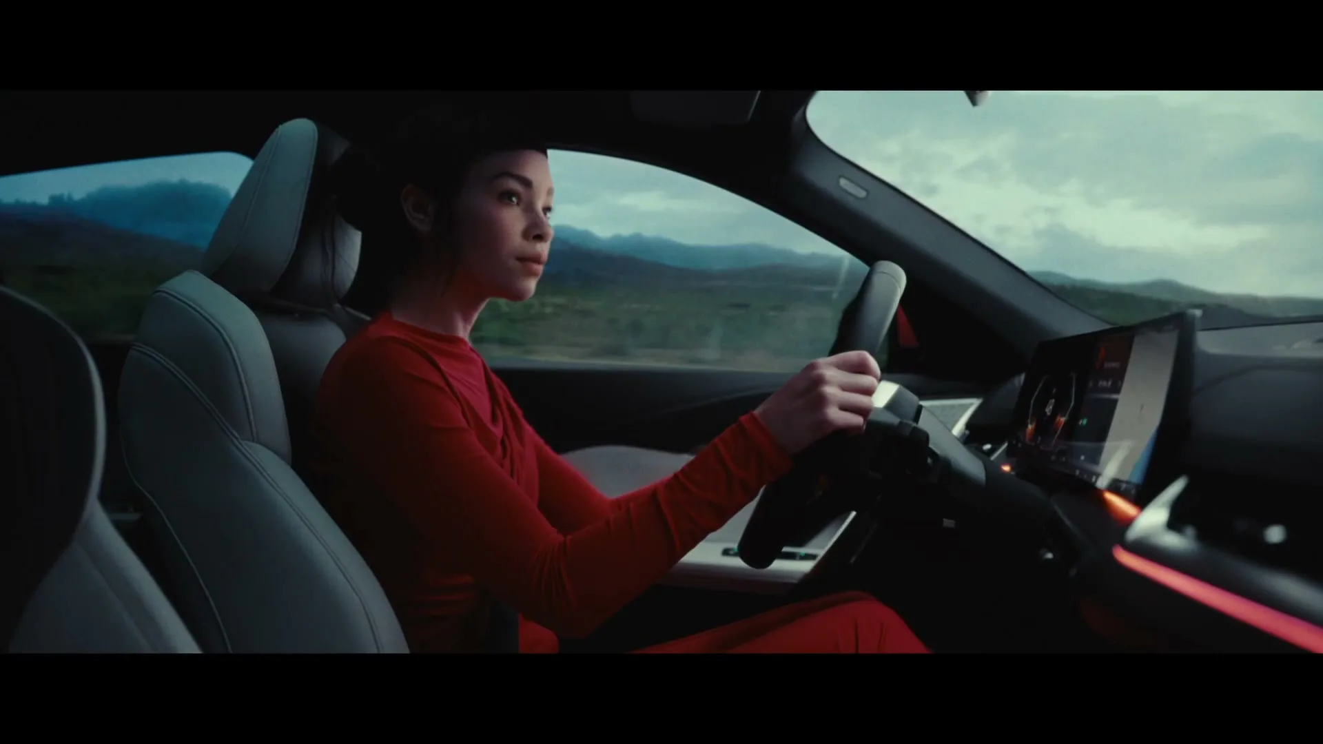 A young woman in a red outfit drives a car, looking forward with a focused expression, as the landscape blurs past outside.