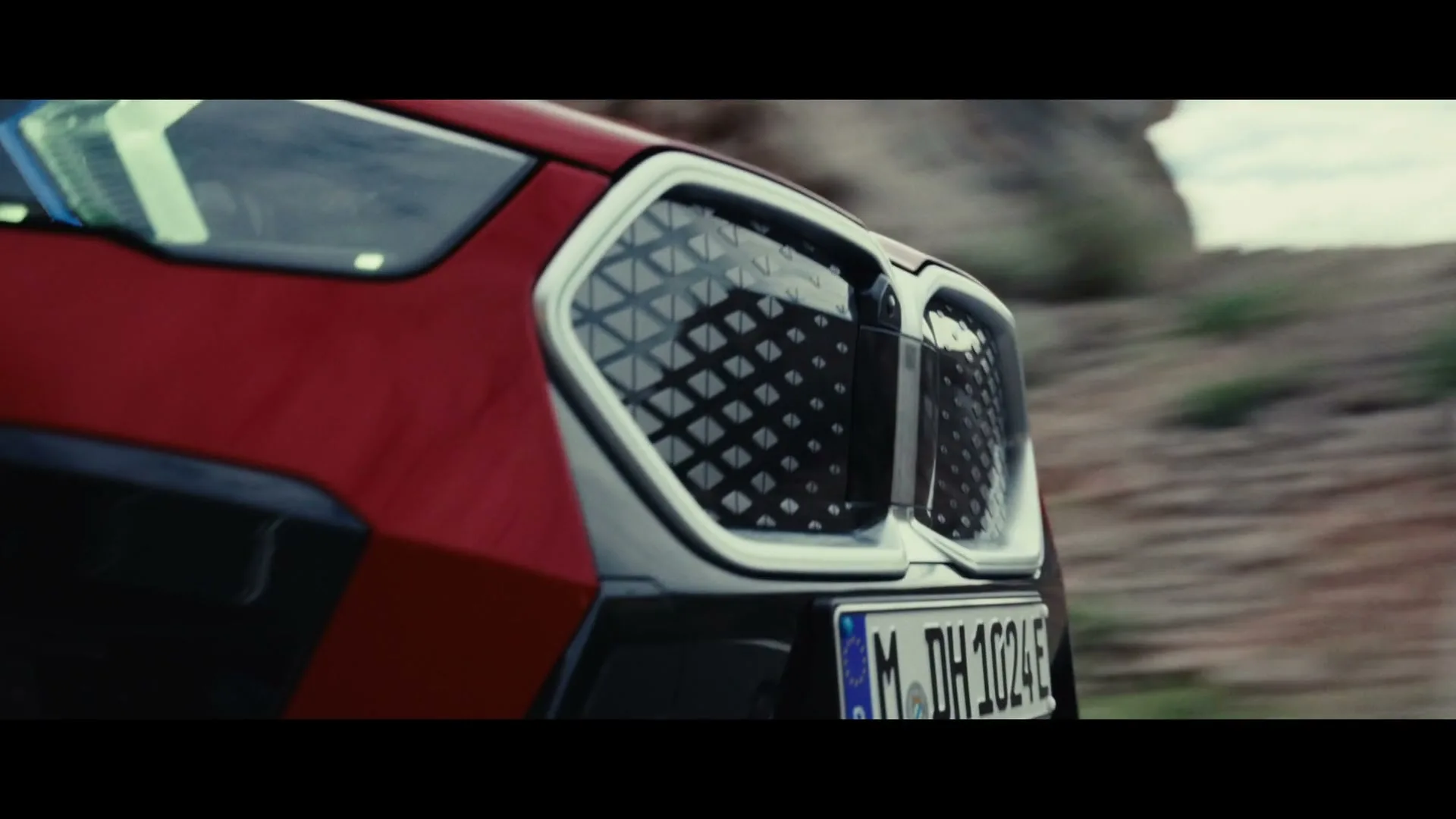 A red BMW car's front grille and headlight are shown in a medium shot, driving through a rocky, outdoor landscape during the day.
