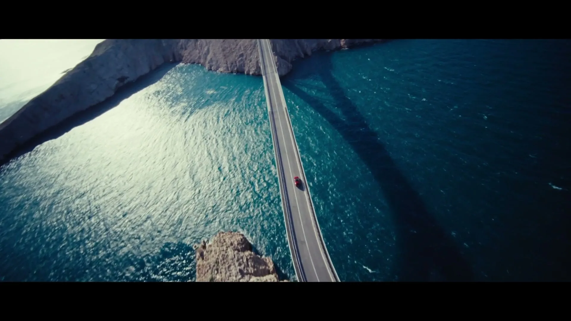 An aerial shot shows a red car driving across a long, narrow bridge spanning a wide body of water with rocky shores under a bright sky.