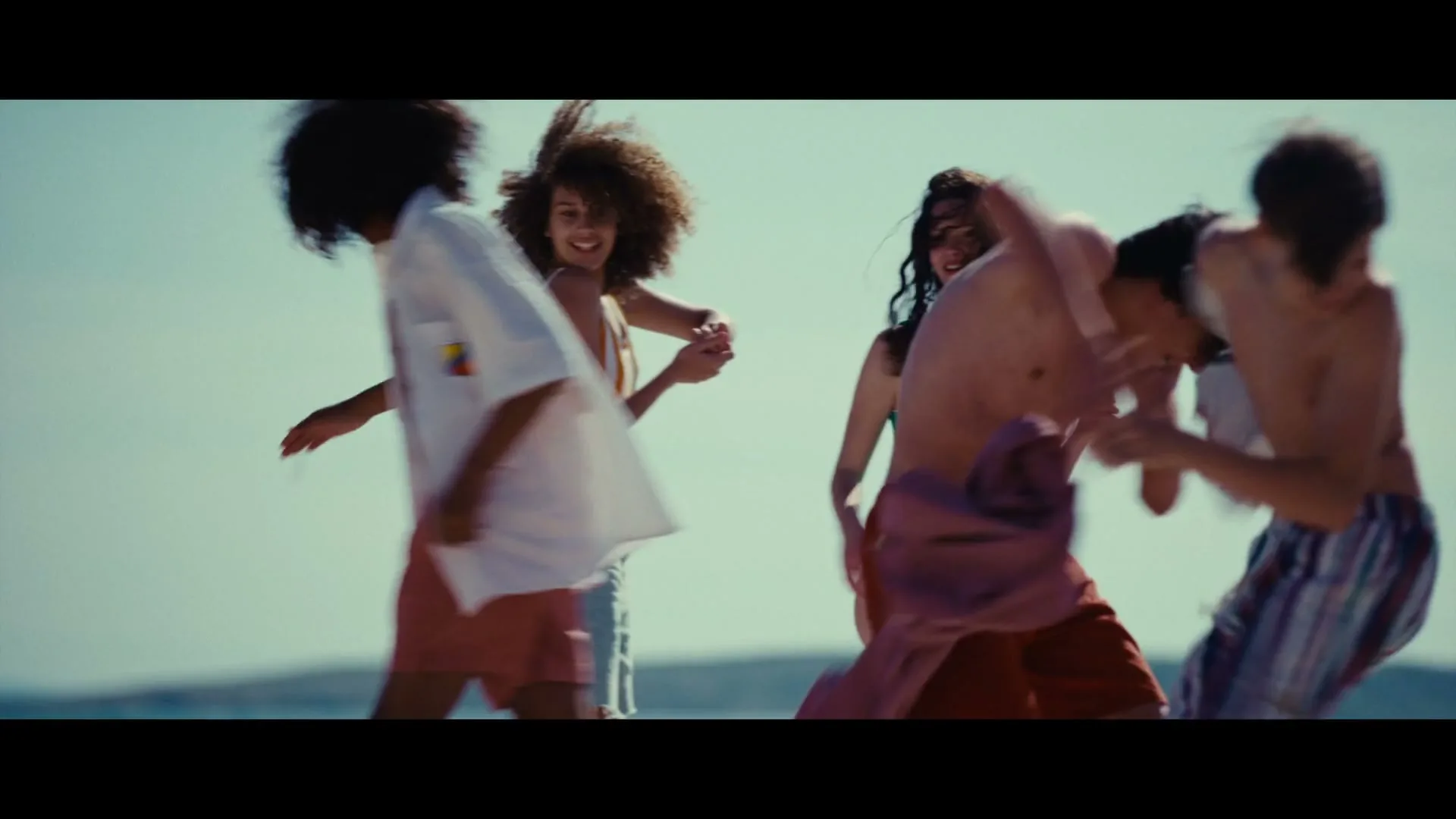 A diverse group of young adults, some in swimwear, are joyfully dancing and laughing on a sunny beach.