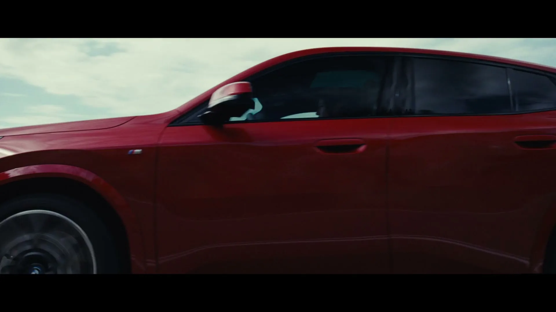 A red BMW SUV drives quickly from left to right on a road under a partly cloudy sky, with motion blur indicating speed.