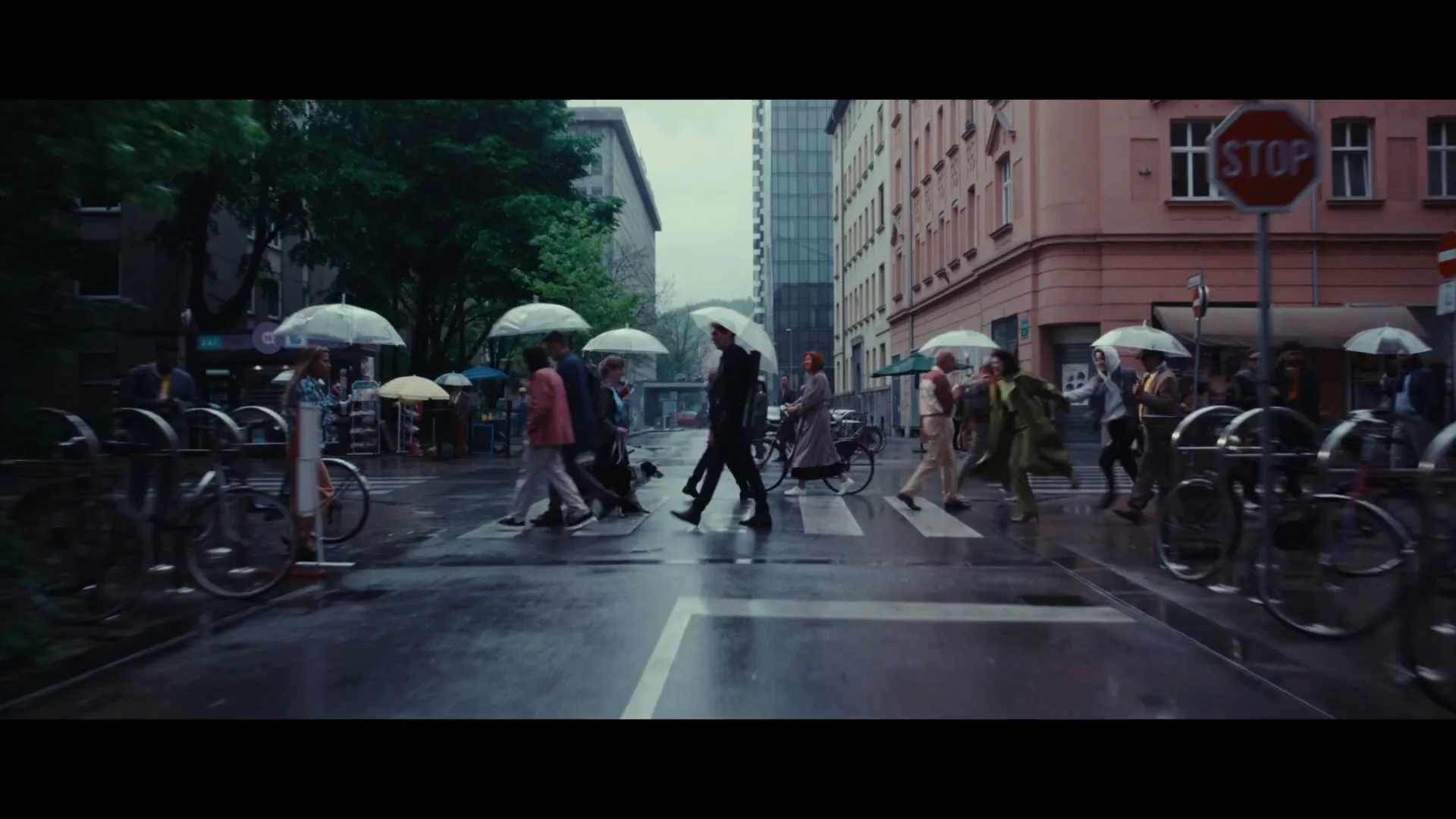 A diverse group of people, dressed in various outfits, walk across a wet crosswalk in a city on a rainy day, holding umbrellas.