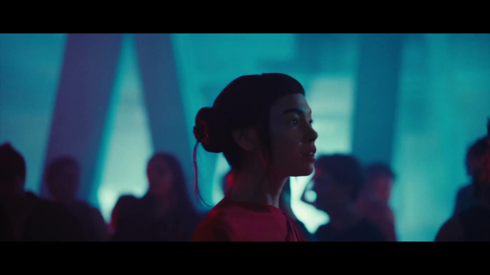 A woman with dark hair in a bun and bangs, wearing a red top, looks to the right in a dimly lit, smoky club with other people in the background.