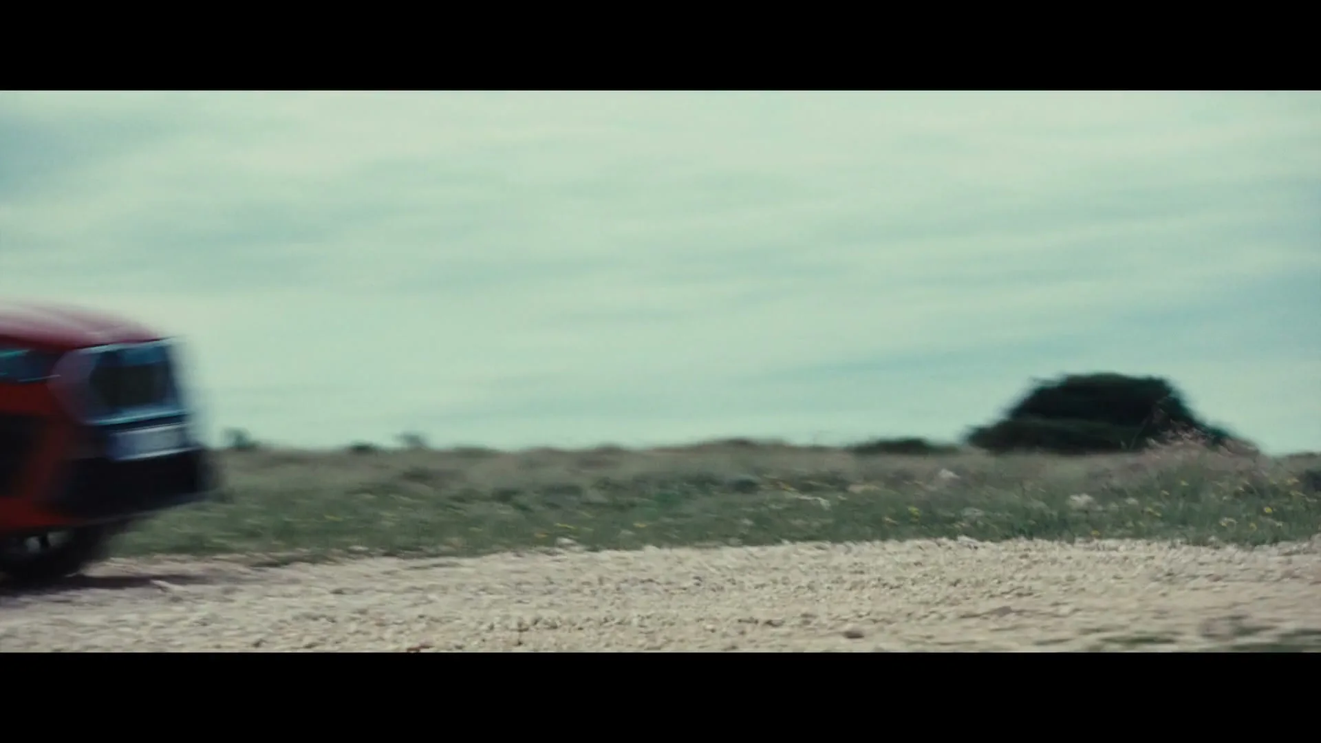 A red BMW car speeds across a dirt road, with green grass and a cloudy sky in the background, creating a sense of motion.