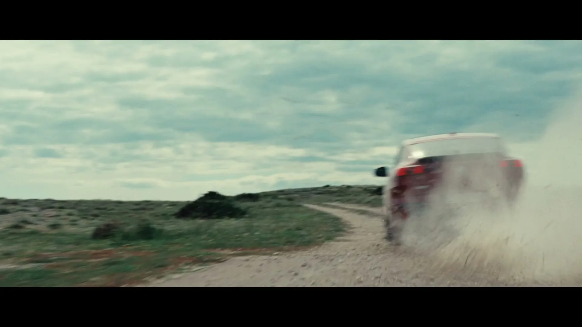 A red SUV drives away on a dirt road, kicking up dust, under a cloudy sky in a rural landscape.