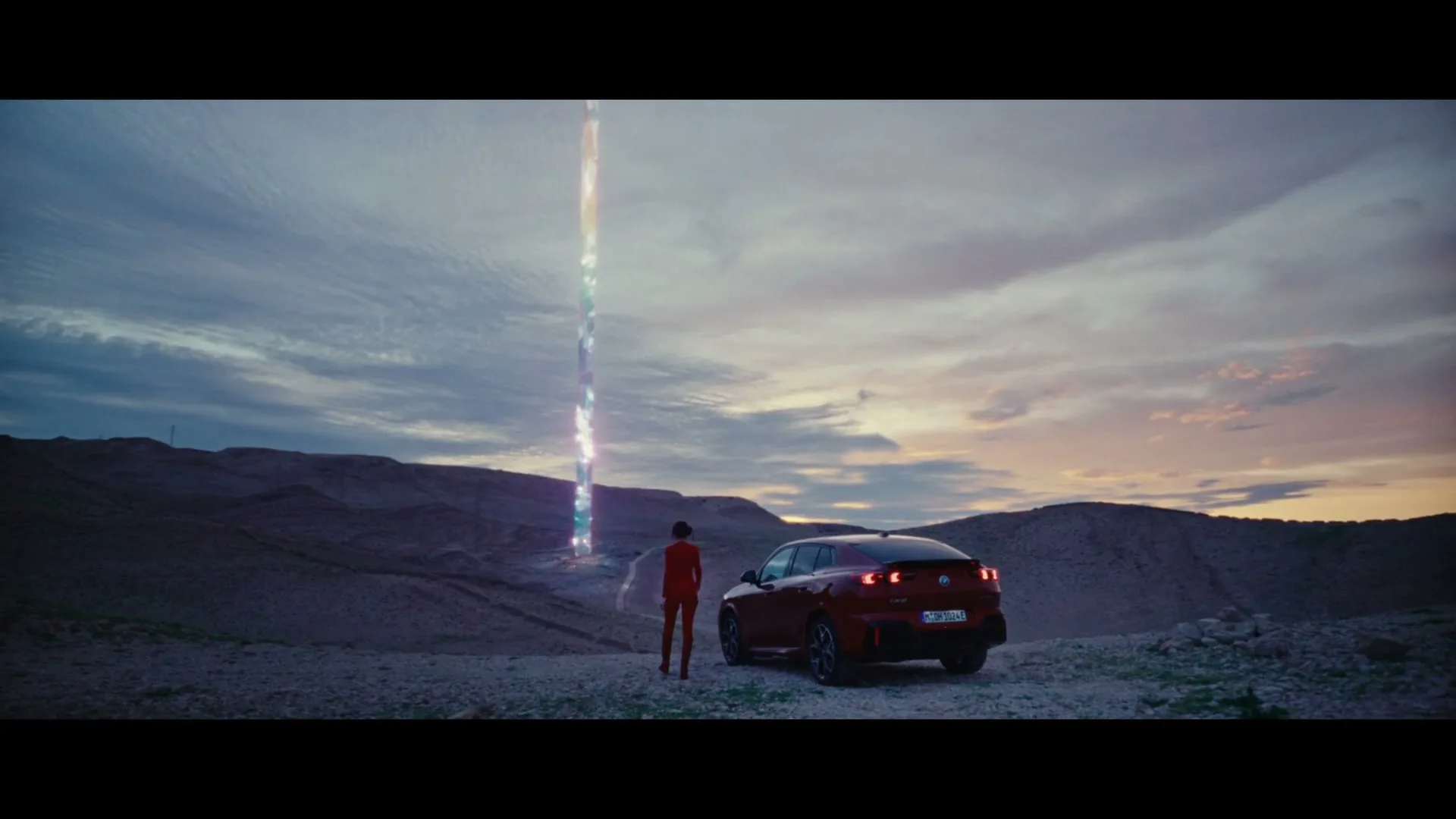 A person in a red jumpsuit stands next to a red car in a desert landscape, looking at a glowing, colorful beam of light rising into the sky.