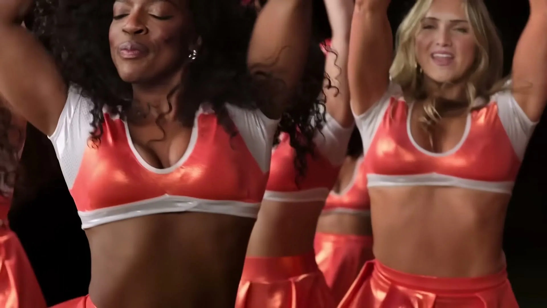 A group of diverse cheerleaders in red and white uniforms dance energetically with their arms raised, smiling and moving in unison against a dark background.