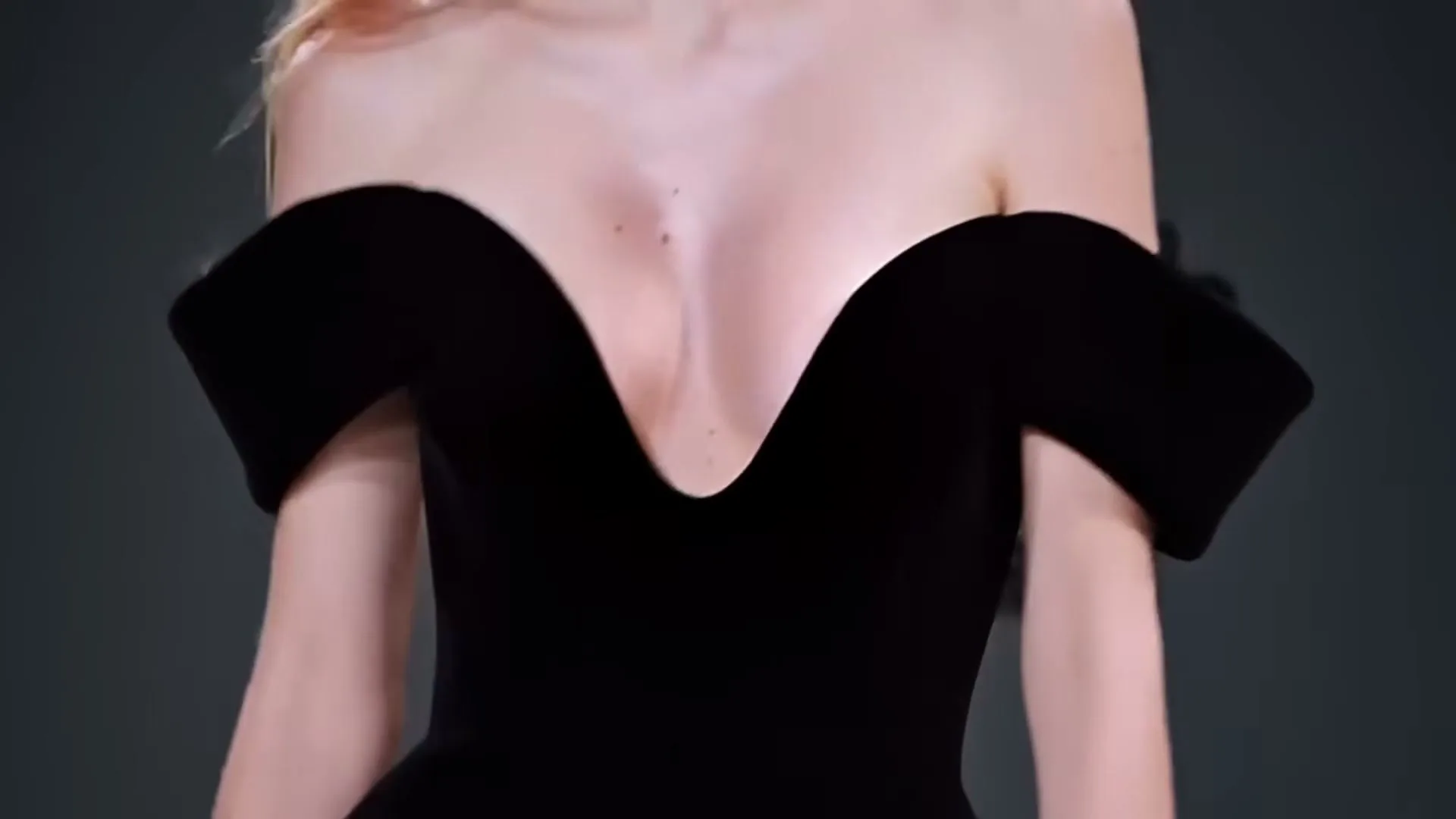 A woman in a black off-the-shoulder dress is shown from the chest up, walking forward in a studio.