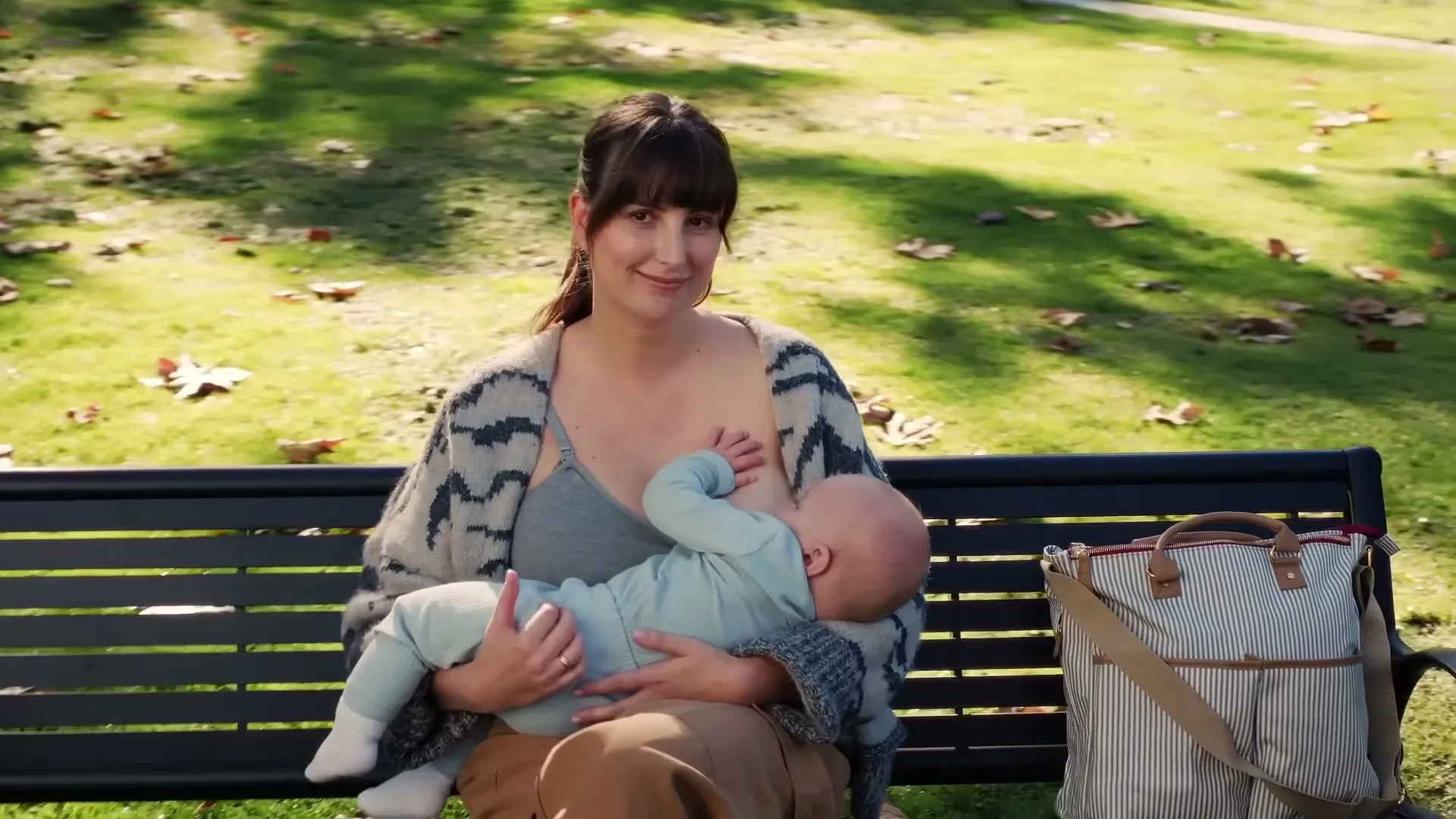 A young mother with dark hair sits on a park bench, breastfeeding her baby, with a diaper bag next to her on a sunny day.