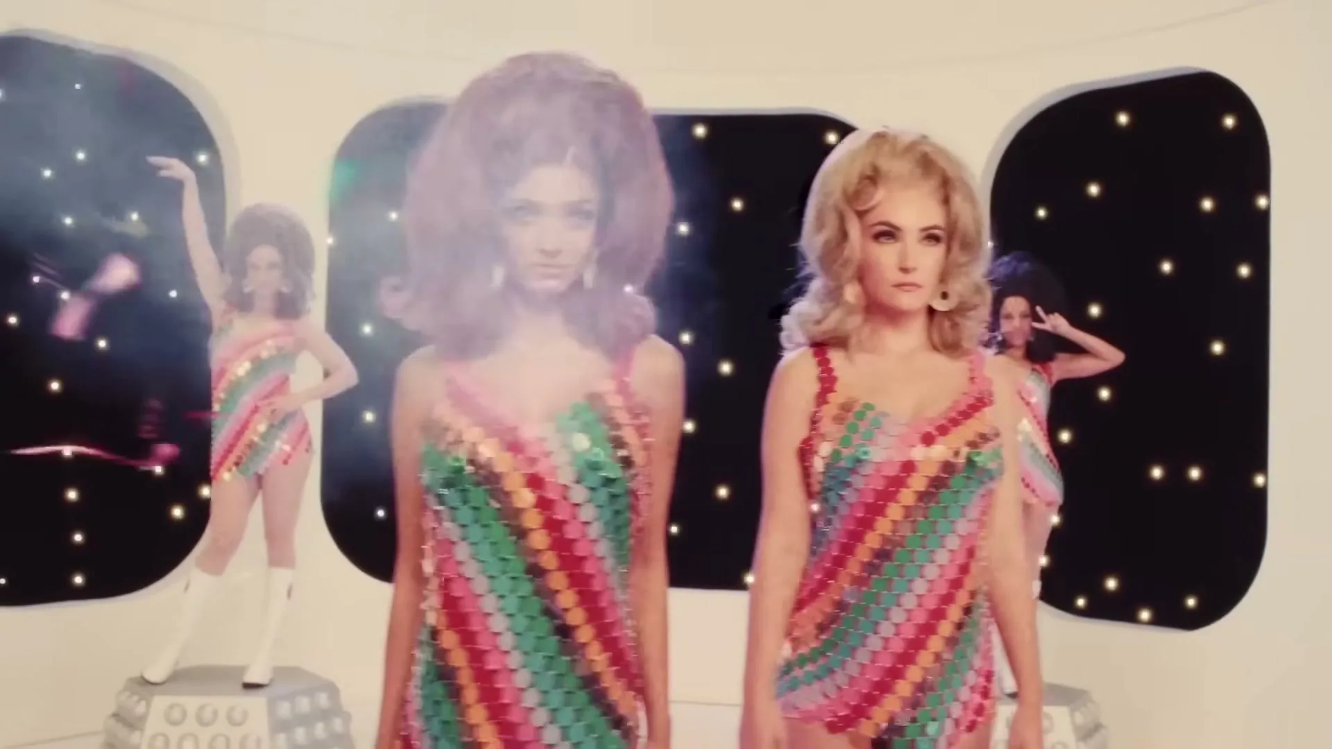 Two women in colorful sequined outfits and big hair stand in a retro-futuristic white room with star-filled windows, while two other women pose in the background.