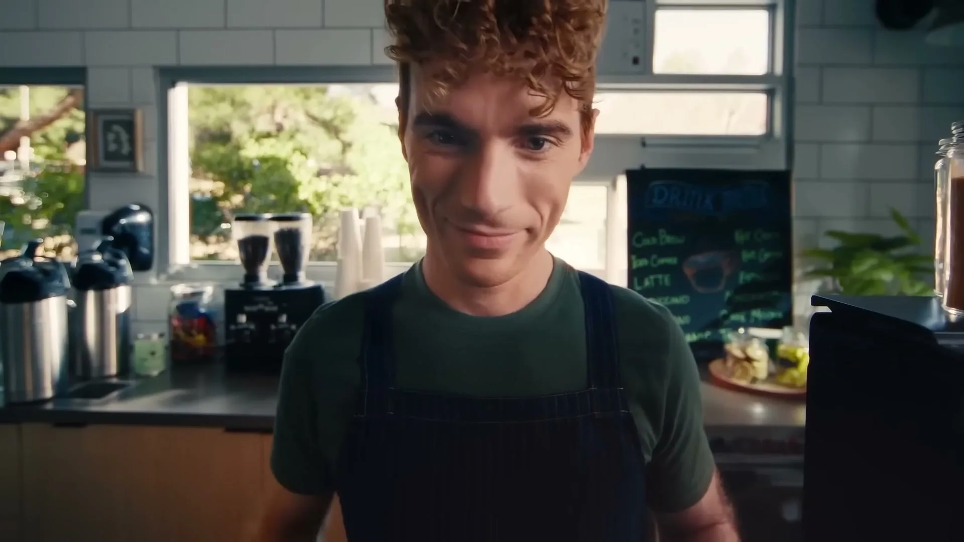 A young male barista with curly red hair and a green shirt smiles mischievously at the camera in a bright cafe.