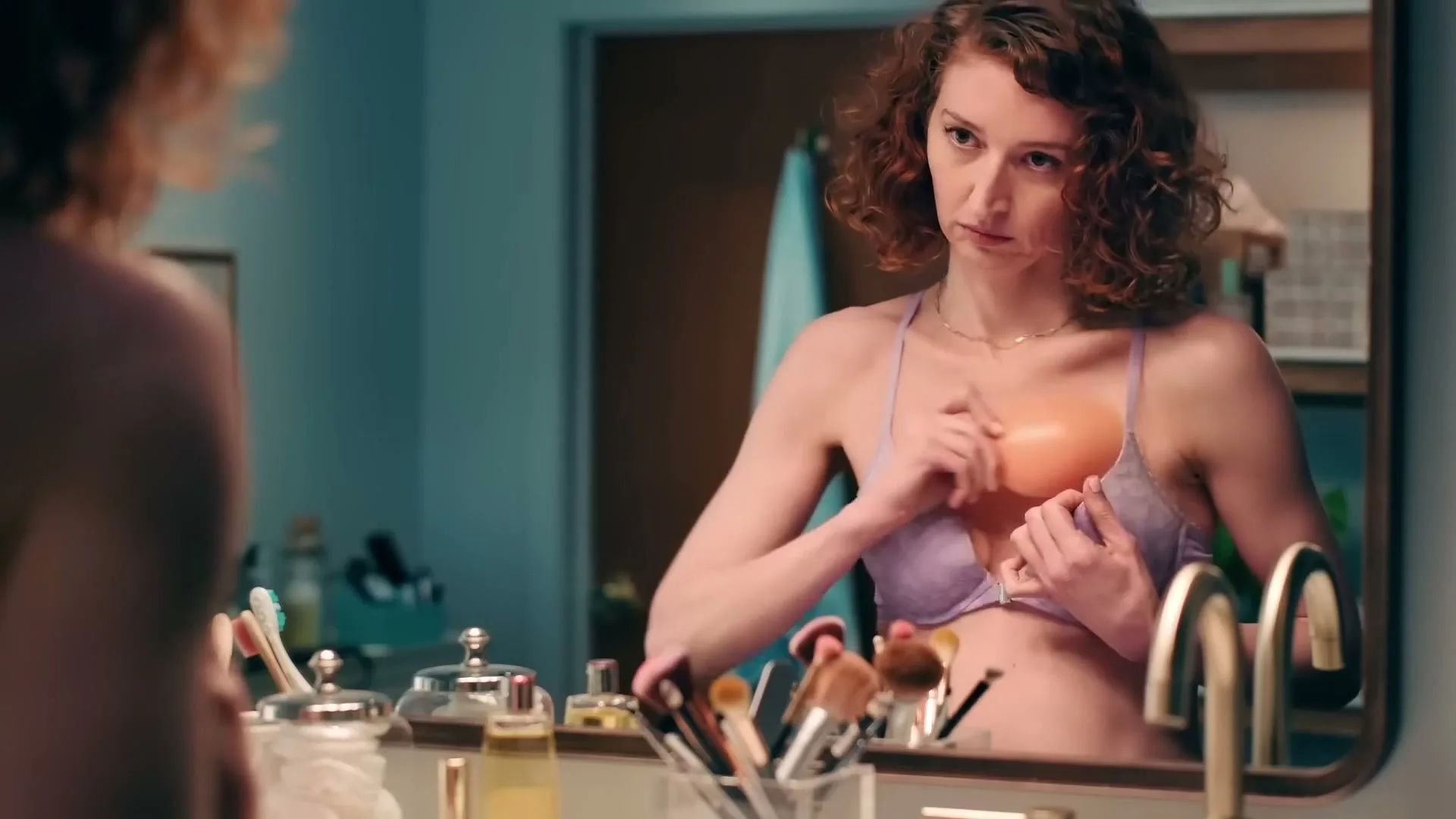 A woman with curly red hair, wearing a lavender bra, looks at herself in a bathroom mirror, adjusting her bra.