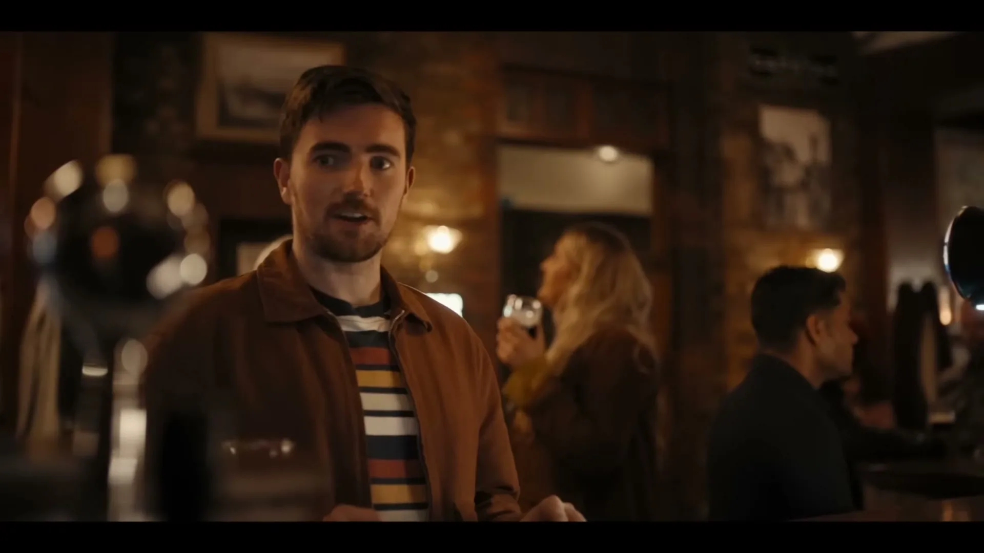 A young man with dark hair and a beard, wearing a brown jacket and striped shirt, stands in a dimly lit pub, looking around and talking.