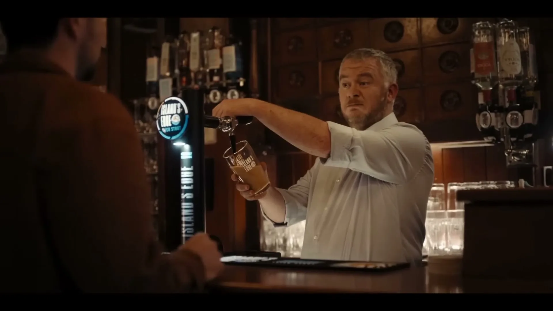 A male bartender with grey hair and a white shirt pours a pint of stout for a male customer at a dark wooden bar in a pub.