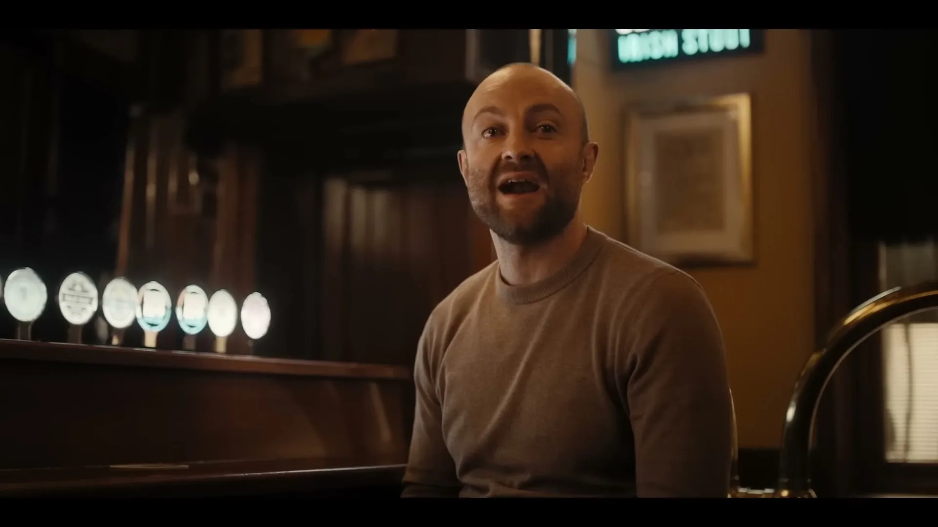 A bald man with a beard sits at a bar, holding a pint of dark beer, and talks to someone off-camera in a dimly lit pub.