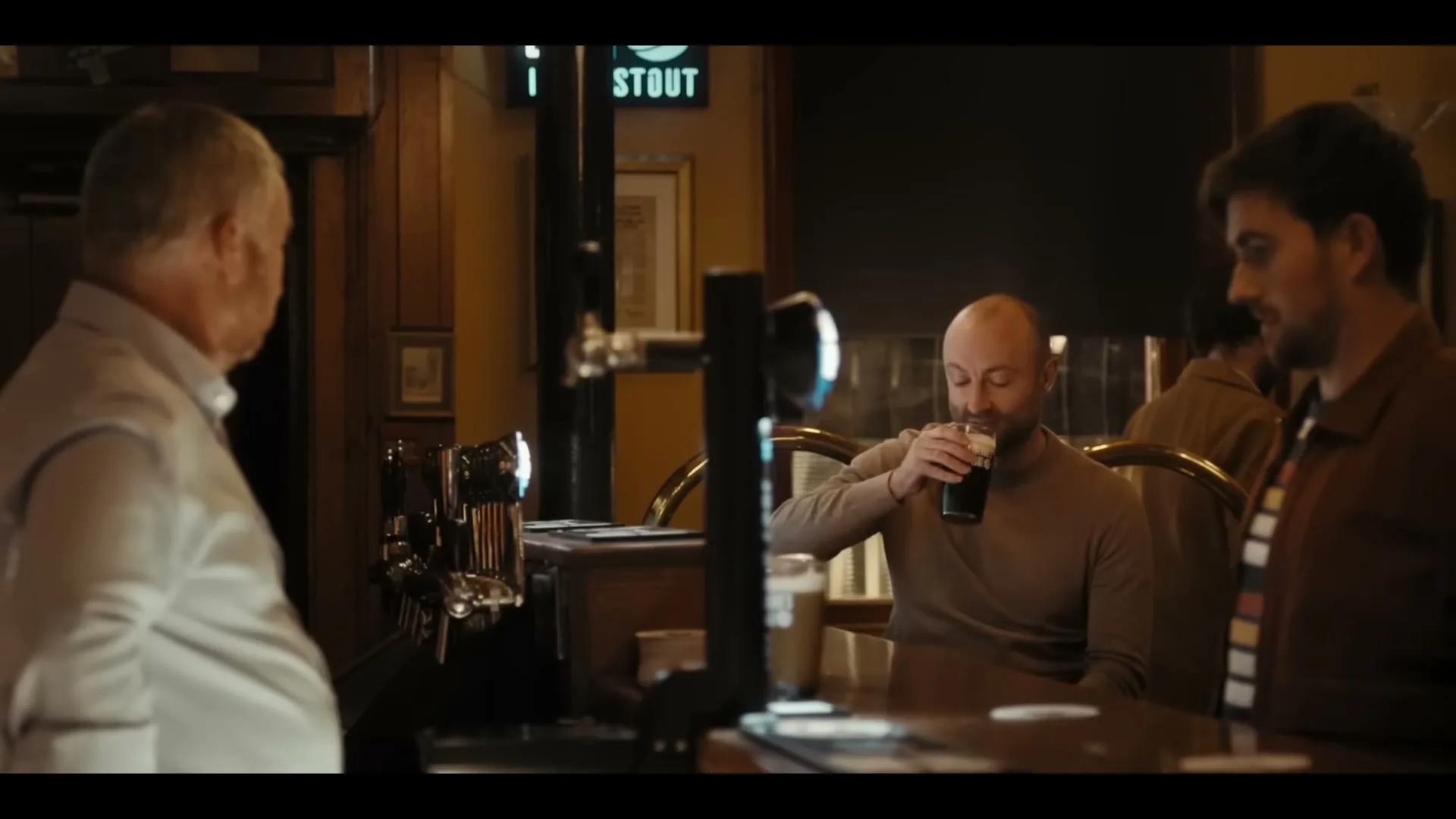 Three men are at a bar, one in a brown shirt takes a sip of his drink, while the other two watch him in a cozy pub.