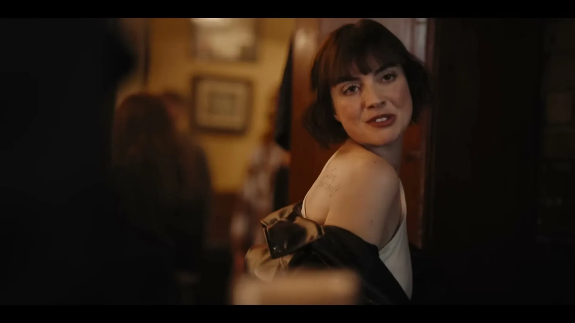 A young woman with short dark hair puts on a black leather jacket in a dimly lit pub, smiling at someone off-camera.