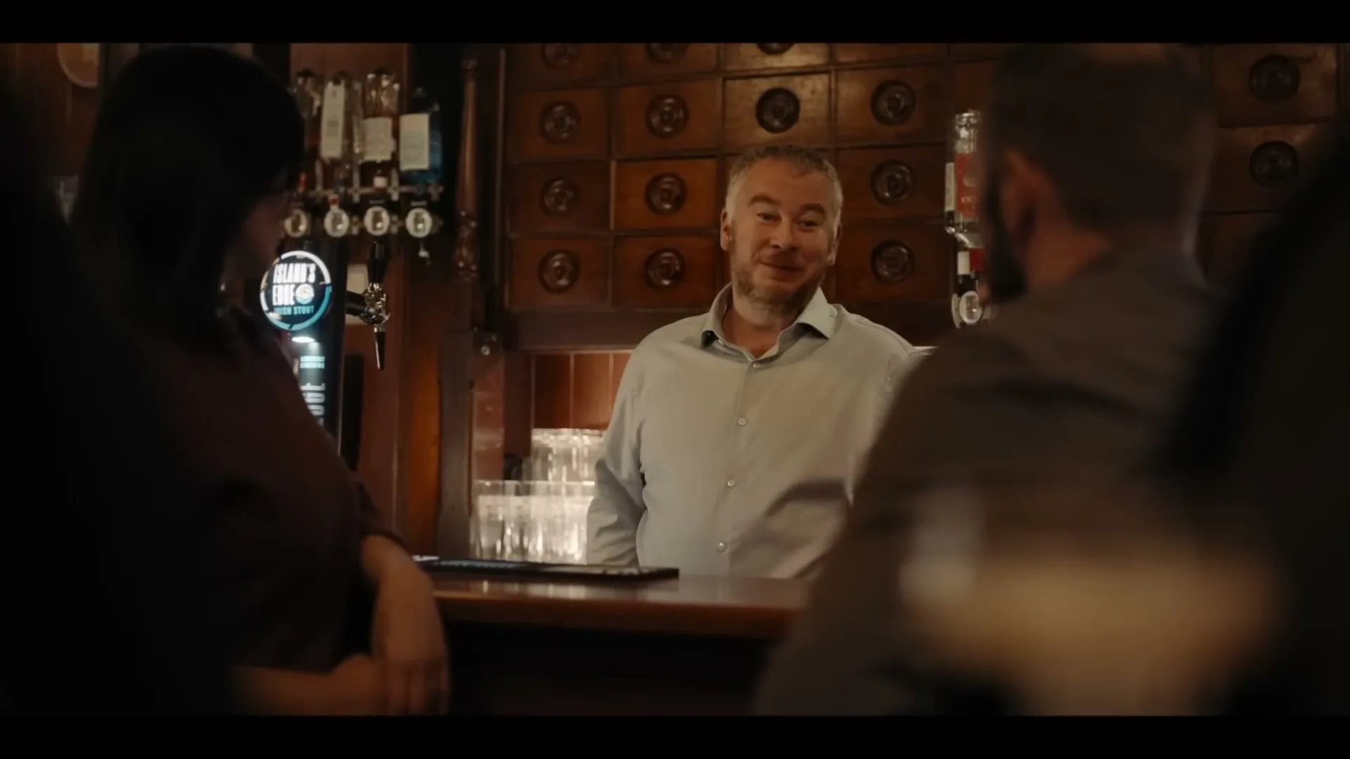 A middle-aged man with grey hair and a light blue shirt stands behind a bar, talking to customers in a dimly lit pub.