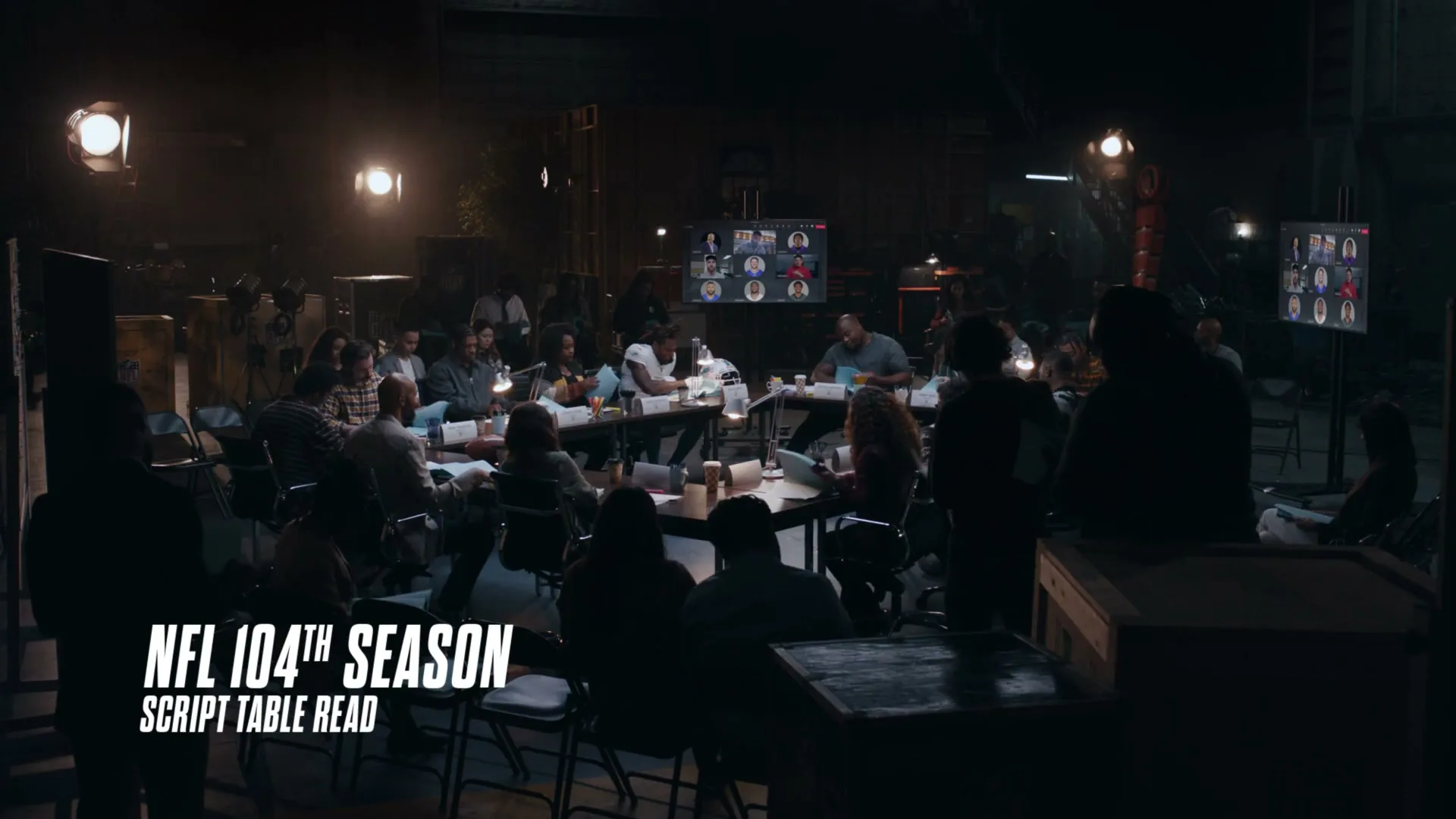 A large group of diverse people sit around tables in a dimly lit studio, participating in a script read for the NFL 104th Season.