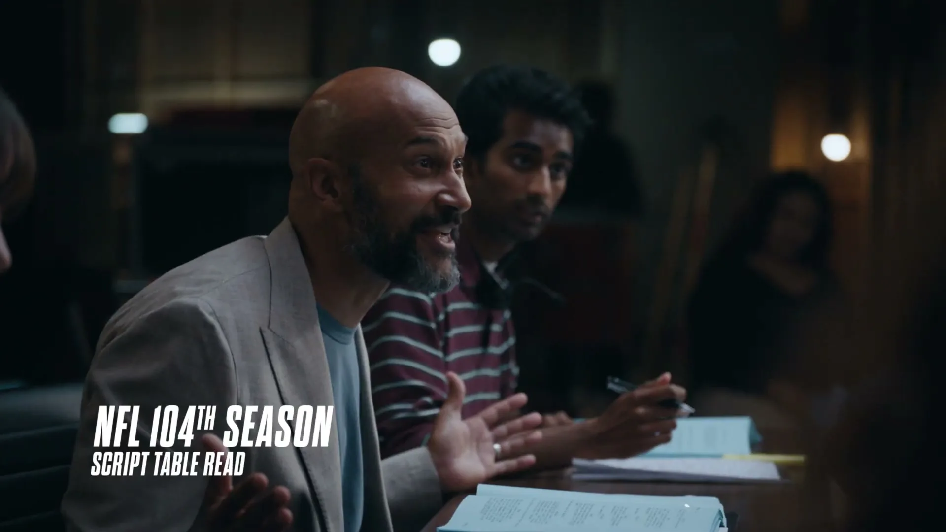 A bald man with a beard, wearing a light jacket, speaks animatedly at a table read, with other people blurred in the background.