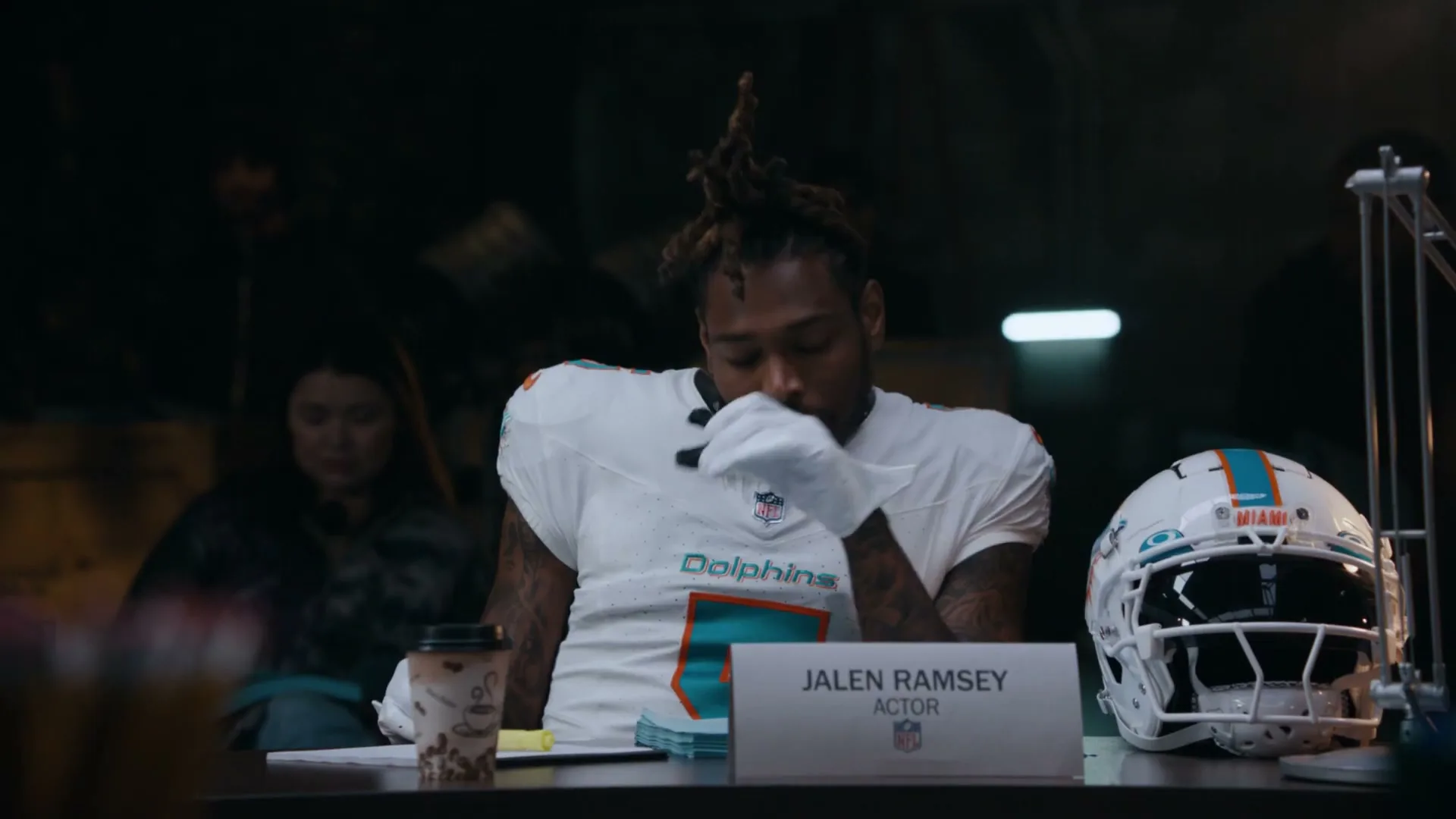 Jalen Ramsey, a male football player in a white Dolphins uniform, sits at a table, looking thoughtful, in a dimly lit room.