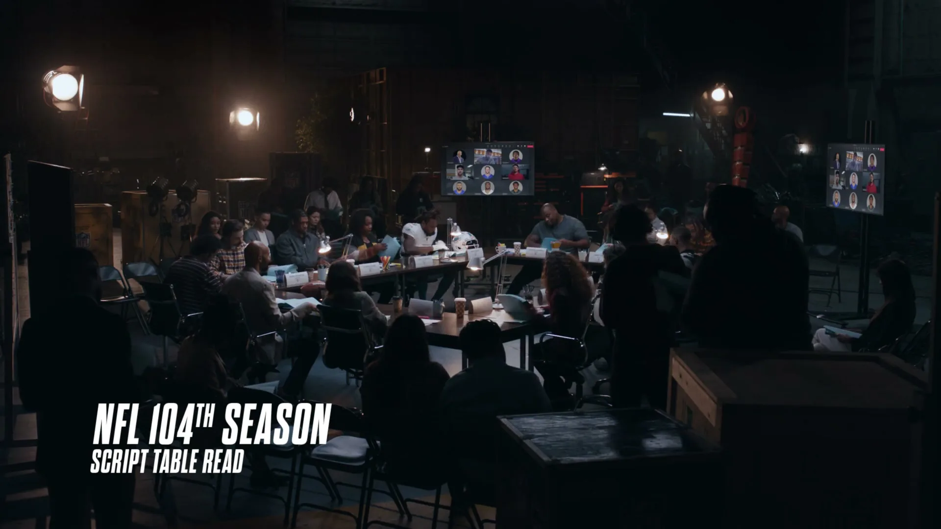 A large group of diverse people sit around tables in a dimly lit studio, participating in a script table read for the NFL 104th Season.