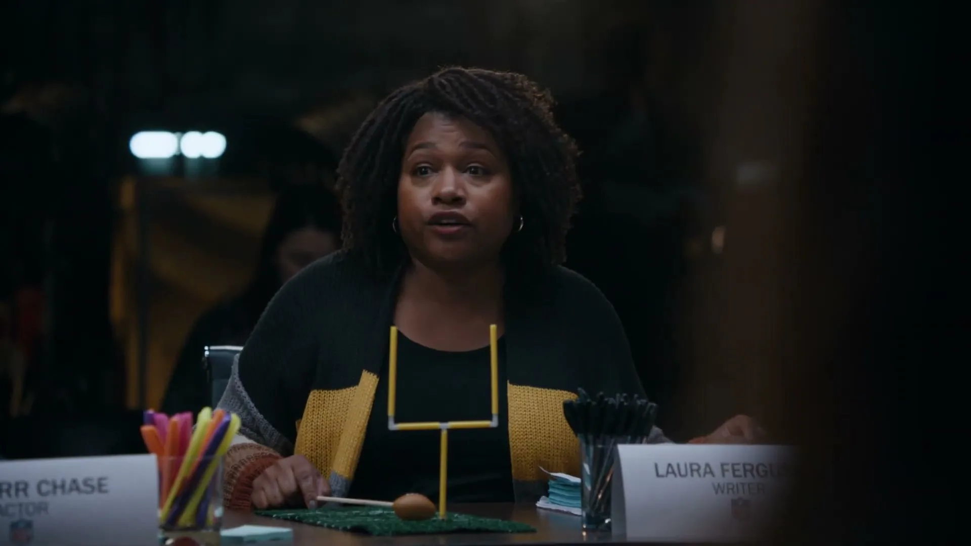 A Black woman with dreadlocks, wearing a black and yellow cardigan, sits at a table, speaking and gesturing with a miniature football and goalpost.