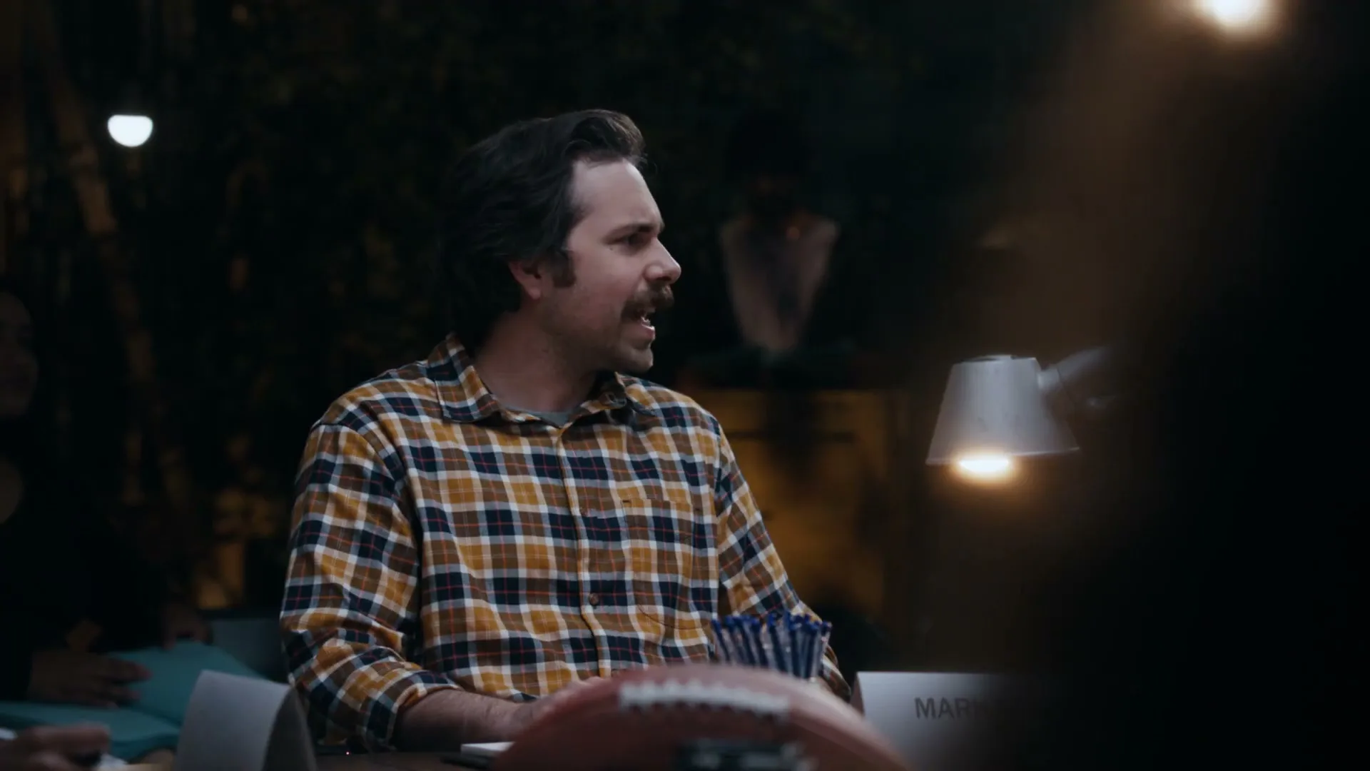A man with a mustache and plaid shirt sits at a table, looking to the right and talking, in a dimly lit outdoor setting at night.