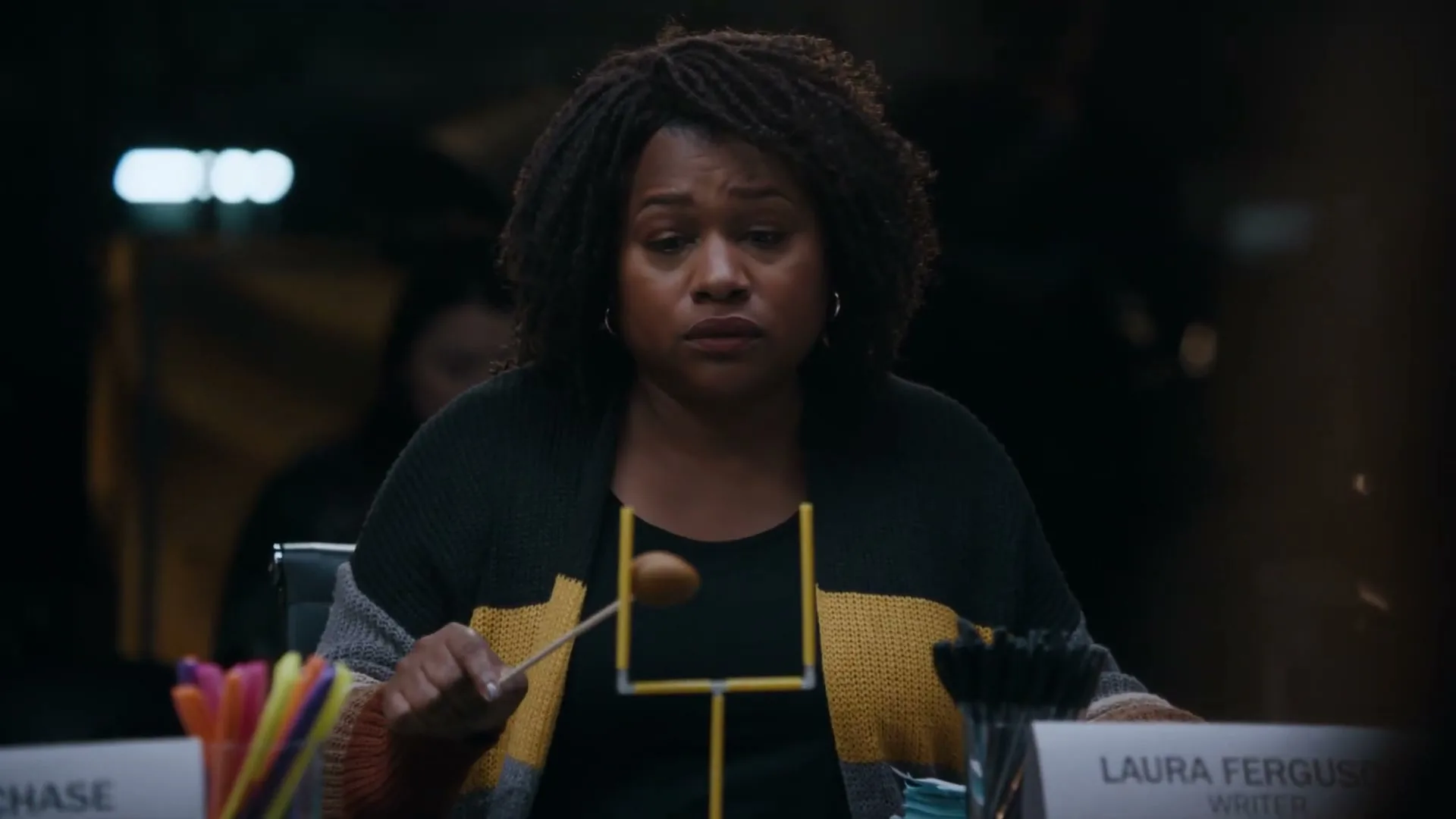 A Black woman with curly hair, wearing a black and yellow cardigan, sits at a table, looking down and playing with a miniature football goalpost and a brown ball on a stick.