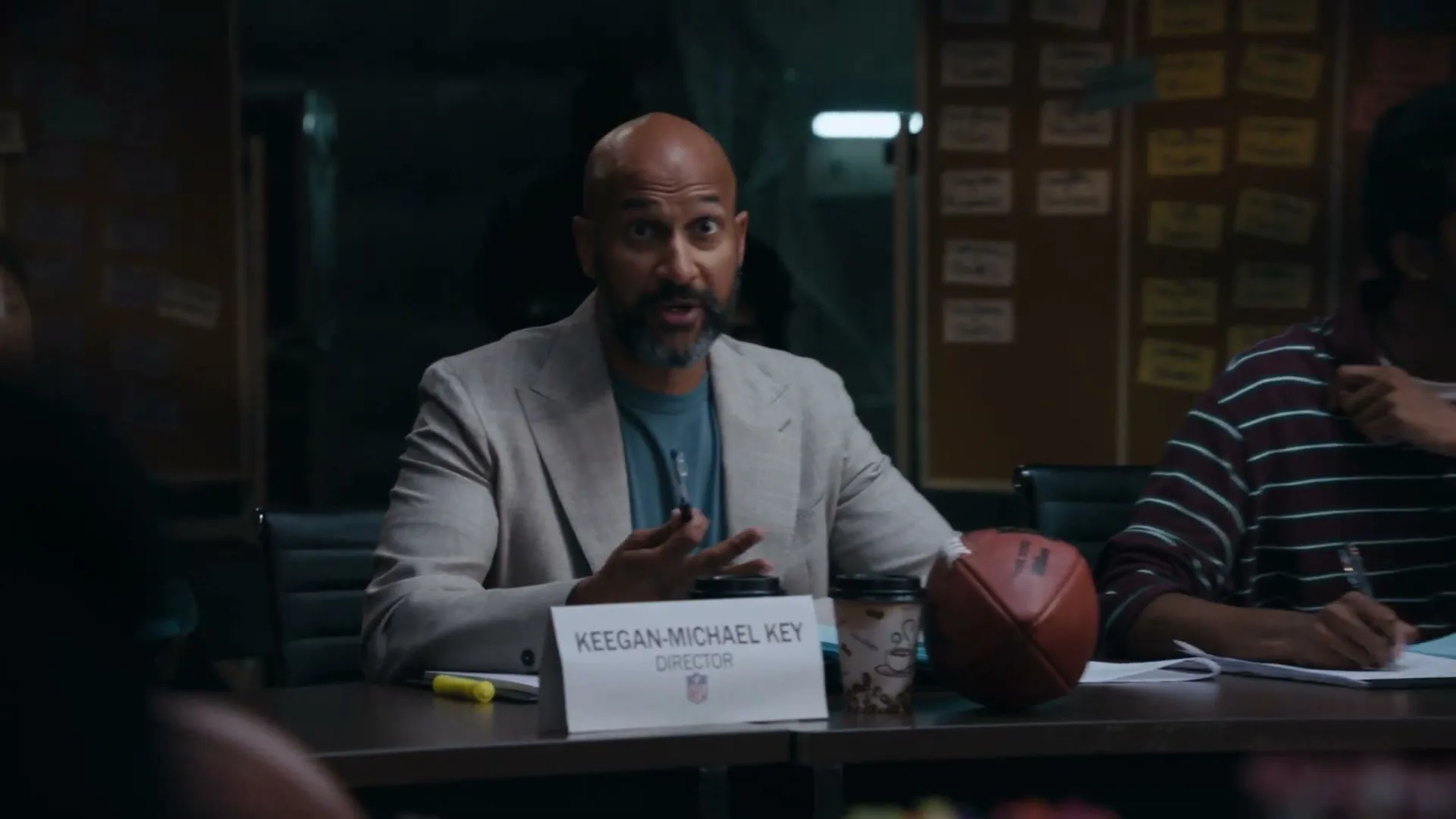 Keegan-Michael Key, a bald man with a beard, wearing a light suit jacket, sits at a conference table, talking and gesturing with his hands.