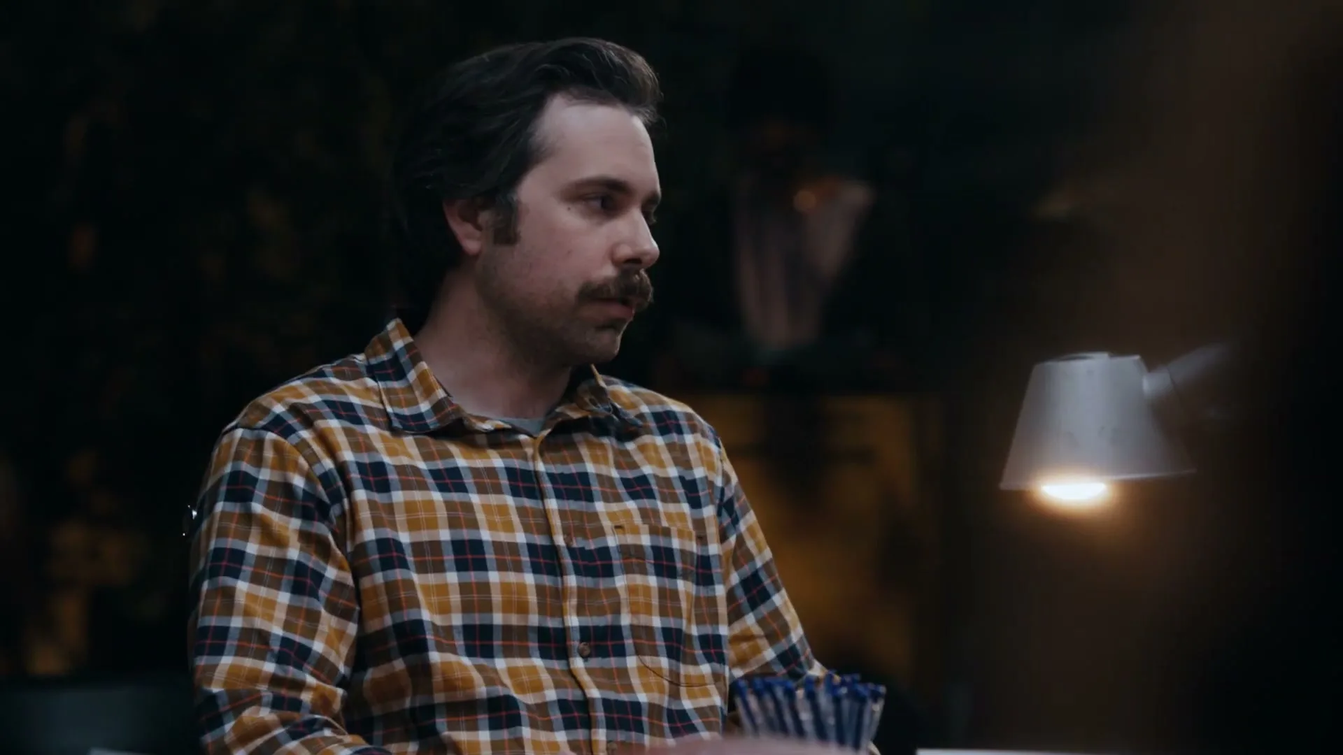 A man with a mustache in a plaid shirt points at a desk lamp, then looks down to write, in a dimly lit room.