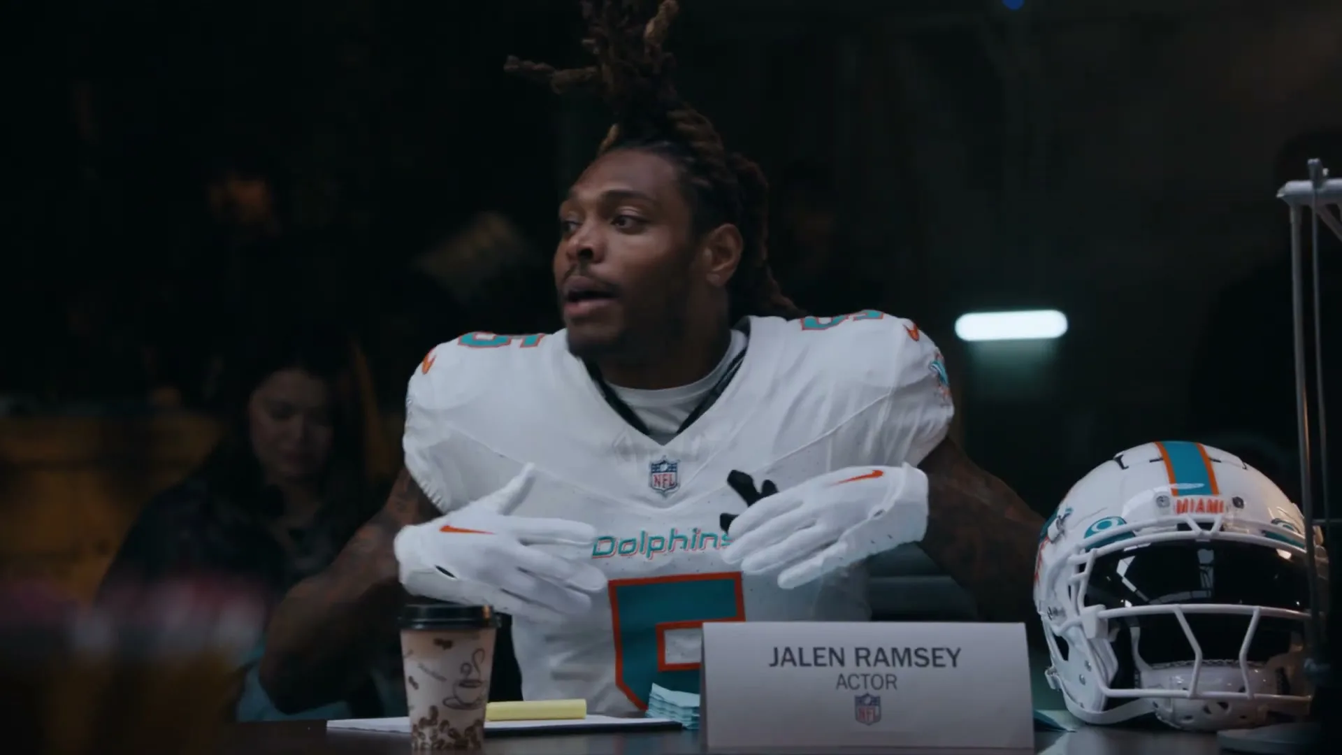 Jalen Ramsey, a male football player in a white Dolphins jersey, sits at a table, gesturing with his hands while speaking in an interior setting.