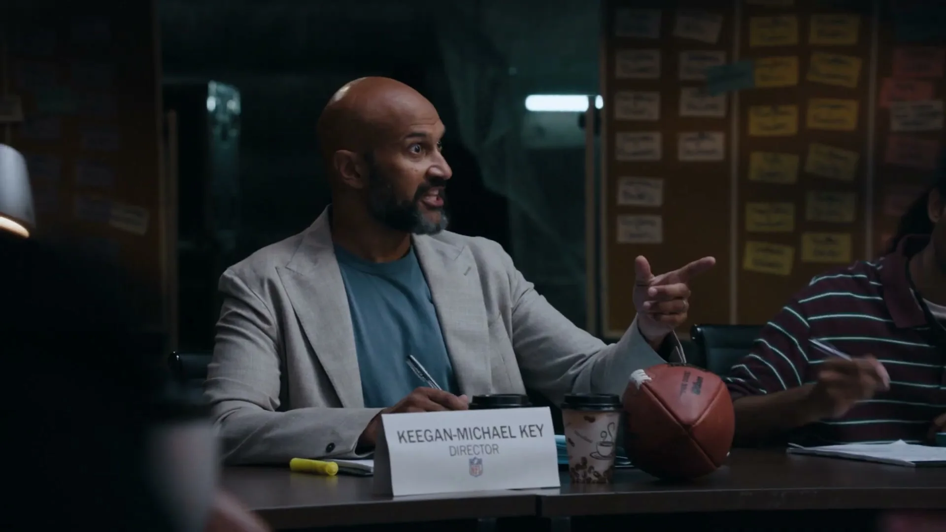 Keegan-Michael Key, a bald man with a beard, wearing a light blazer and blue t-shirt, sits at a conference table, pointing and talking to someone off-screen.