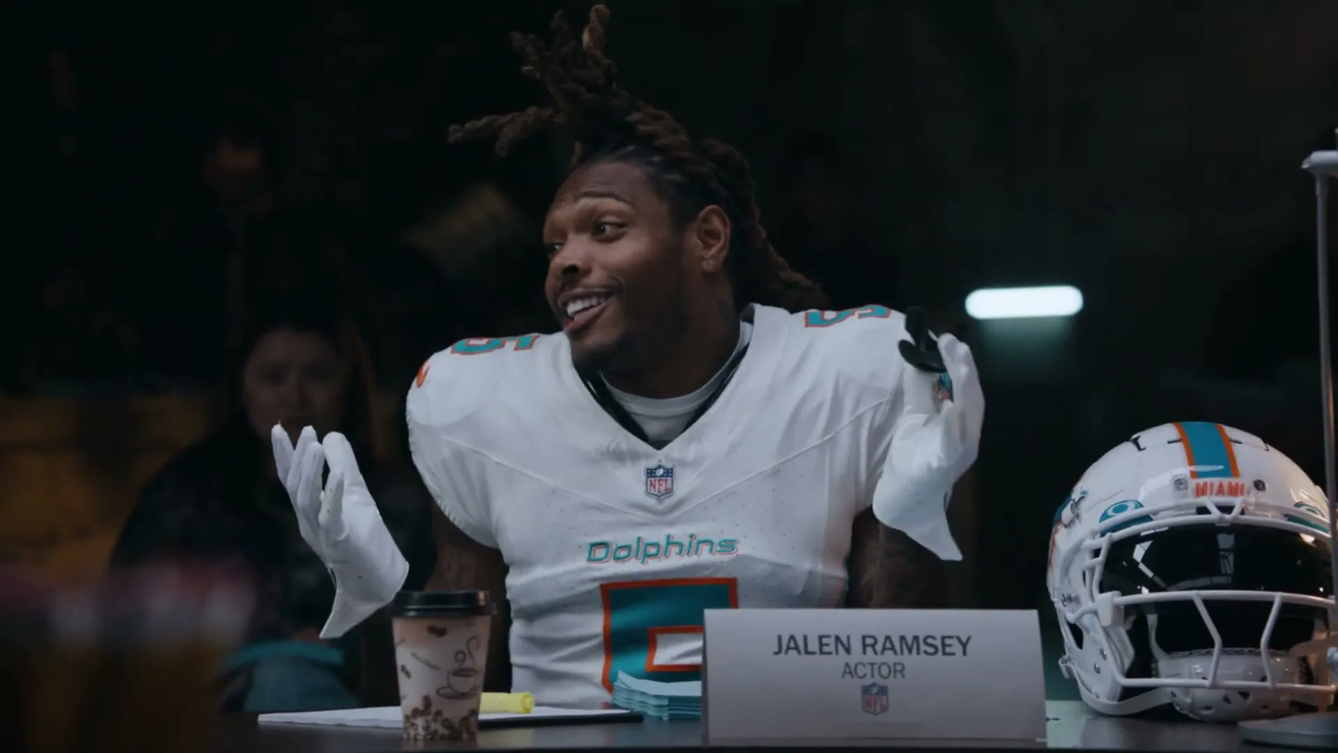 Jalen Ramsey, a male football player in a white Dolphins jersey, sits at a table, shrugging and gesturing with white-gloved hands in an interior setting.